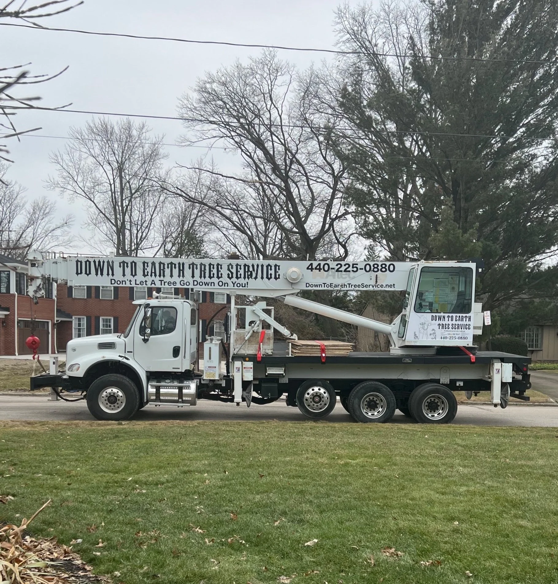 A truck with a crane attachment for tree service, parked on a residential street with houses and leafless trees in the background.