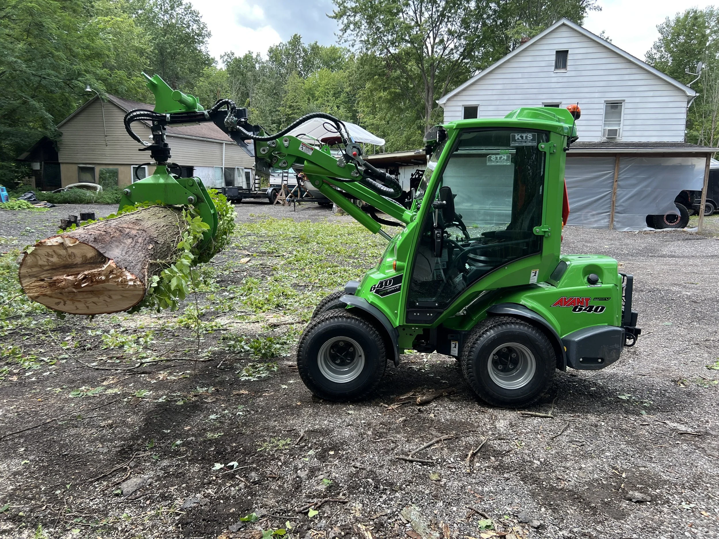 A green compact utility loader lifting a large fallen tree trunk with surrounding buildings and trees in the background.