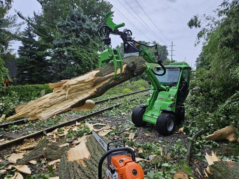 A small green skid-steer loader using a hydraulic grapple attachment to lift and move a large fallen tree trunk on a railway track, with debris and chopped wood scattered around.