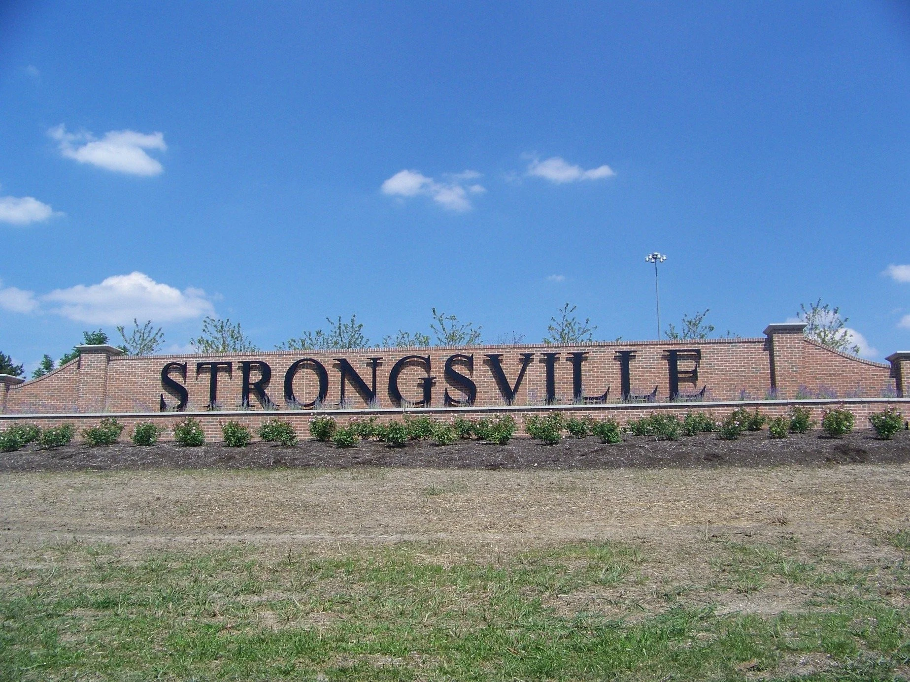 Large brick sign that reads 'Strongsville' in black letters, with a landscaped area and a blue sky with clouds in the background.