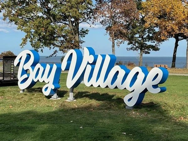 Large three-dimensional sign that reads "Bay Village" in cursive white letters with a blue shadow, set on a grassy area near trees and a body of water in the background.