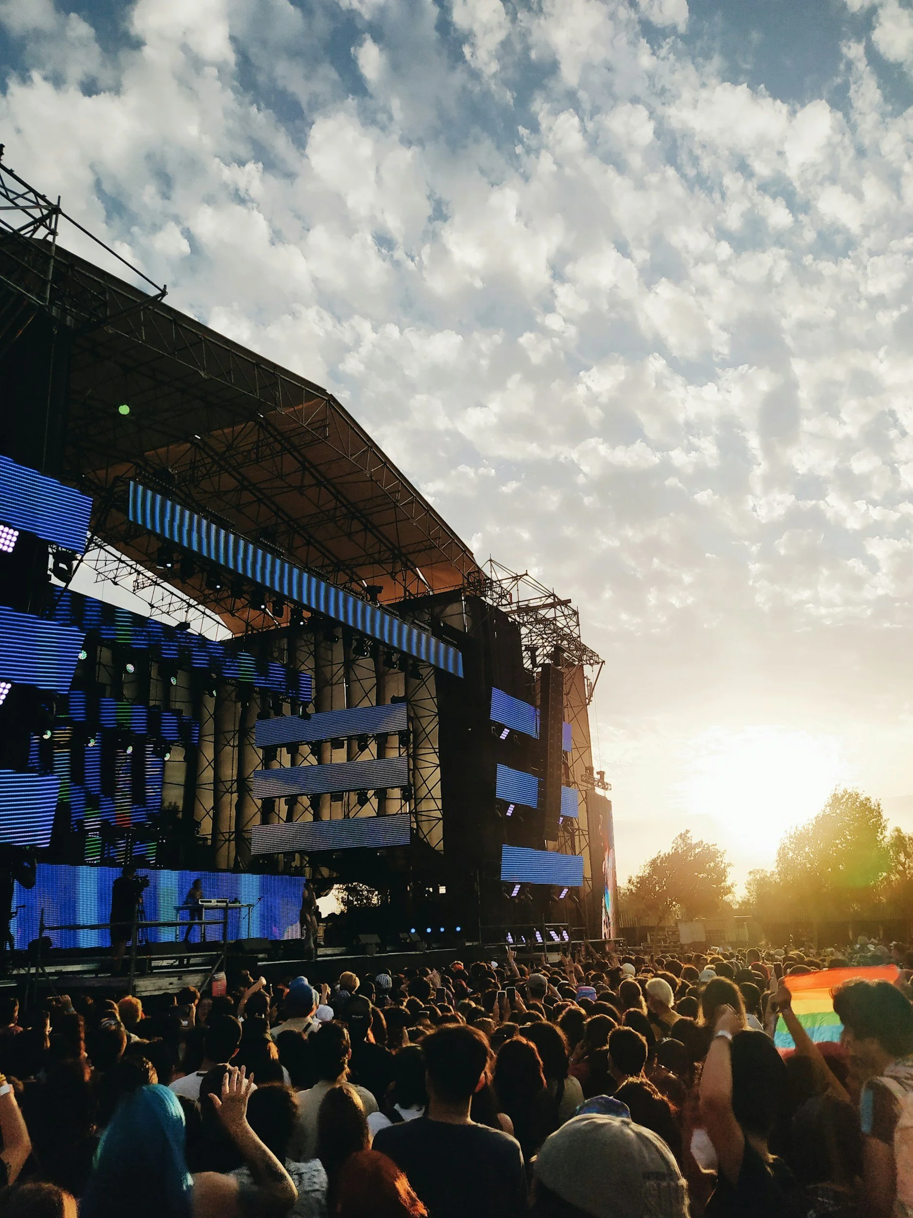 A large outdoor concert stage with digital screens, set against a sky with scattered clouds and the sun setting in the distance. There is a crowd of people watching the concert.