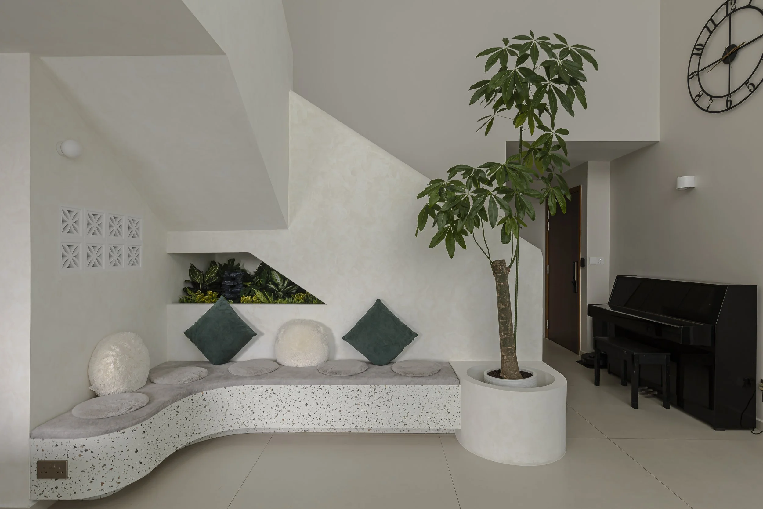 Modern indoor space with white terrazzo bench, green cushions, large potted tree, black piano, avant-garde clock, and decorative elements