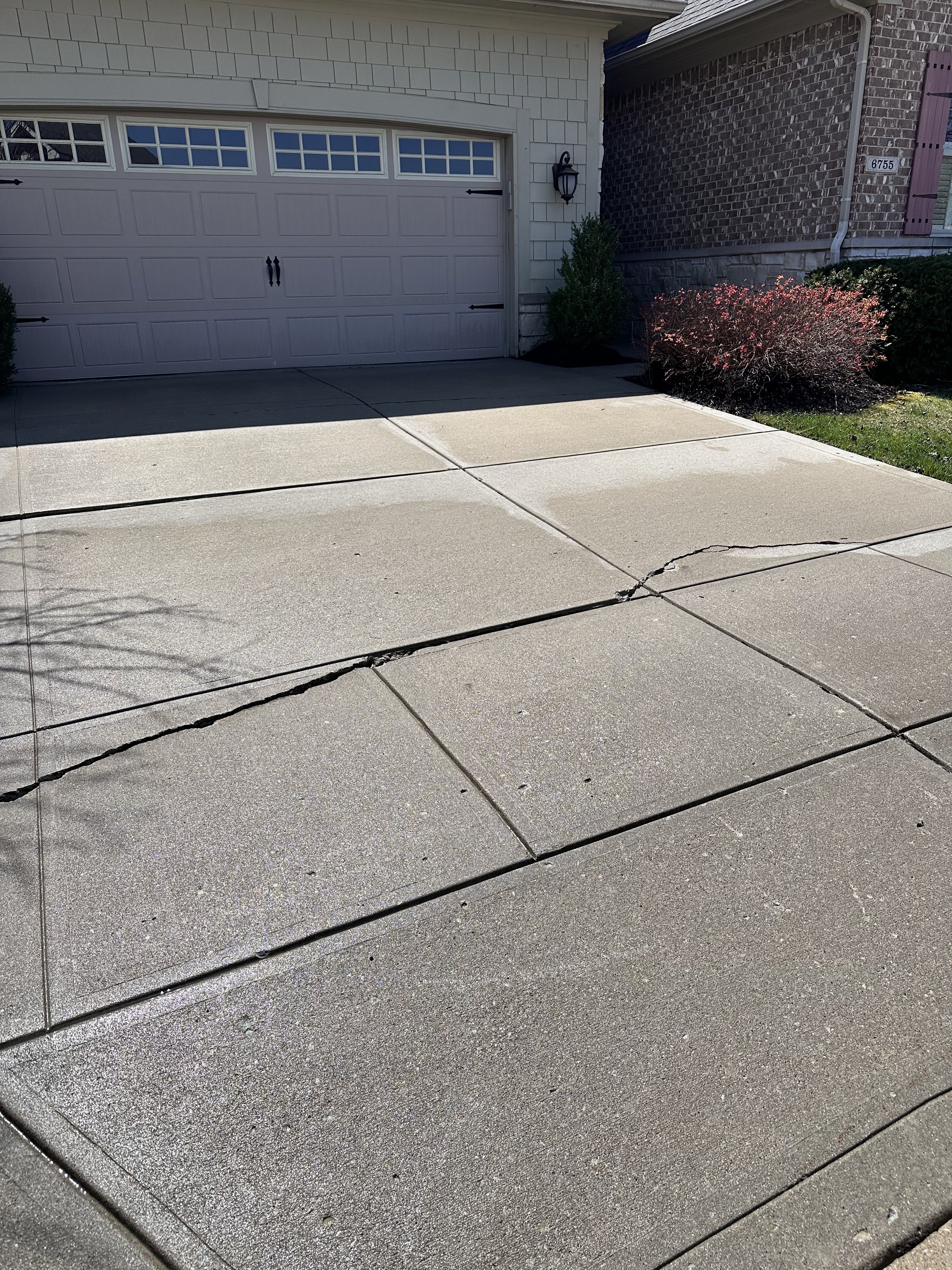 Front view of a residential garage with three-panel door, concrete driveway, and a visible crack. Landscaping includes shrubs and a lawn adjacent to brick exterior walls.
