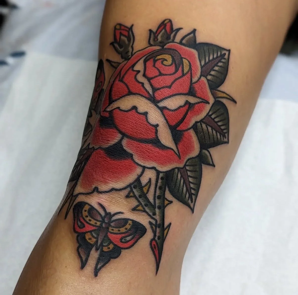 Tatouage d'une rose rouge avec des feuilles vertes et un papillon jaune et noir en dessous.