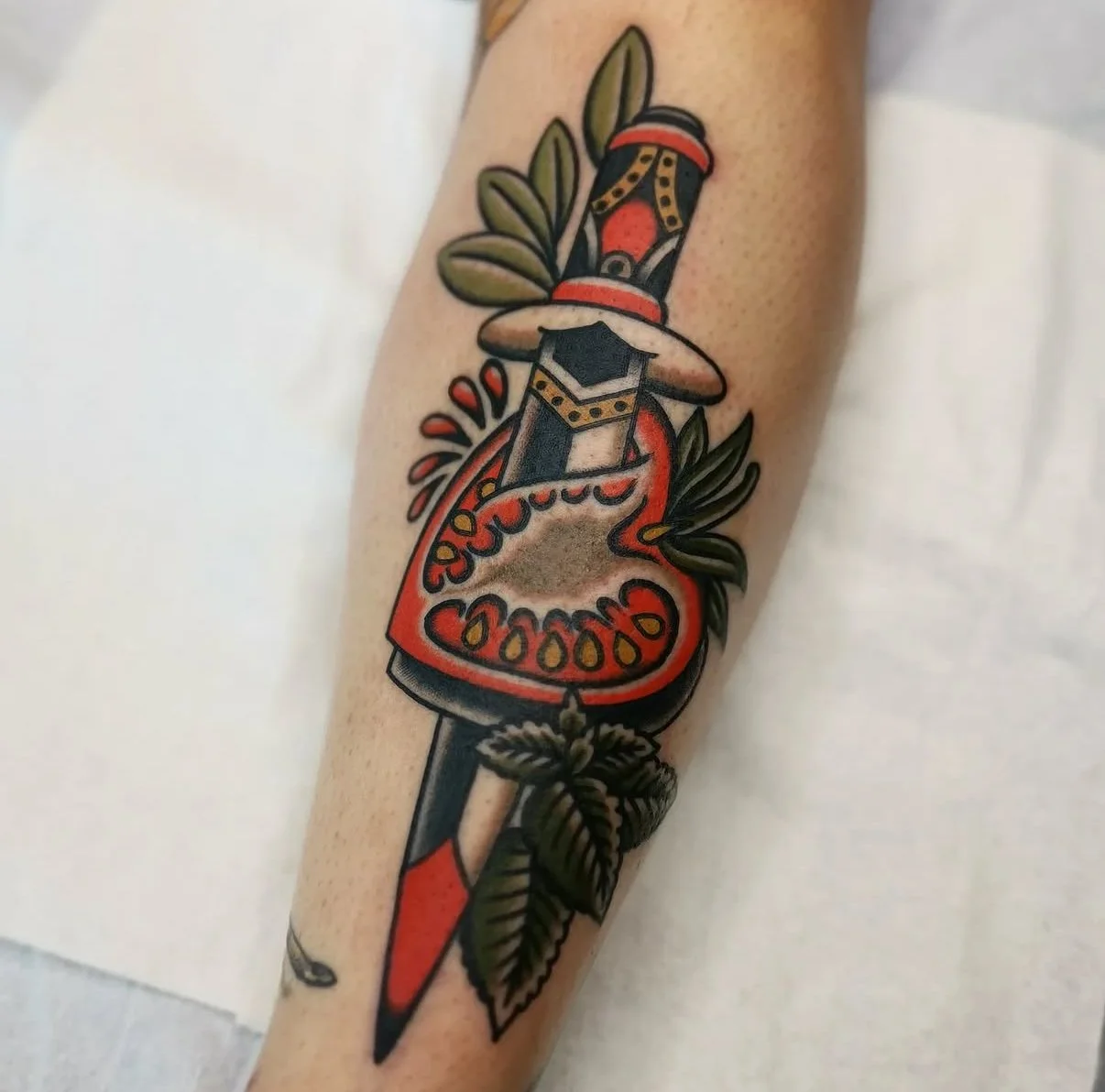 Tatouage d'une pomme coupée en deux avec un couteau, des feuilles et un couteau noir, rouge, vert et beige.