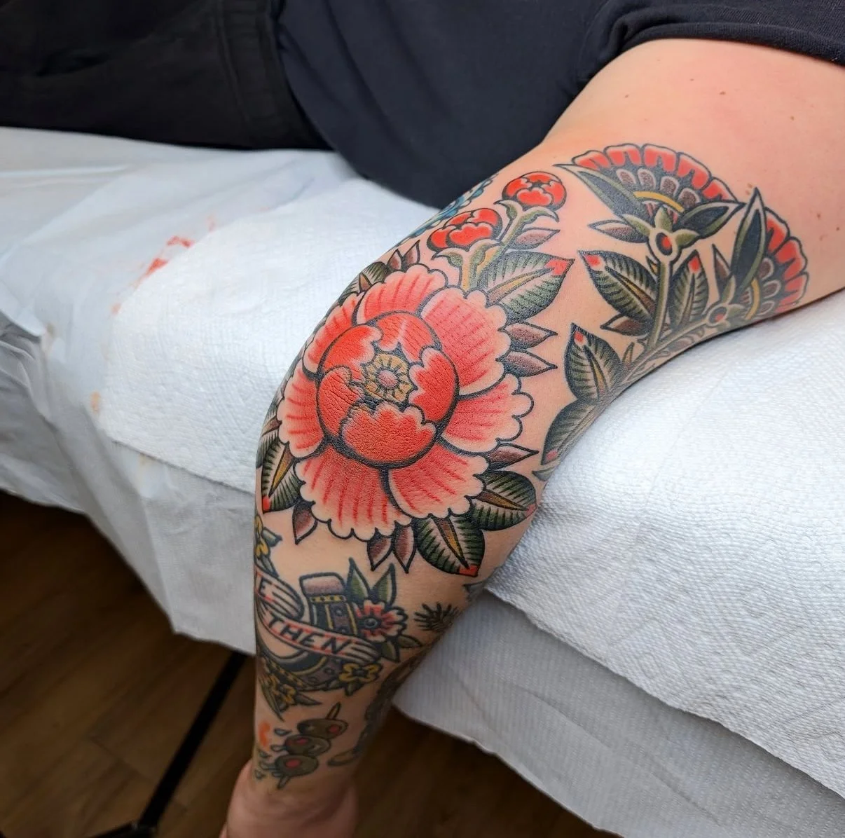 Un bras tatoué avec un grand tatouage floral de pivoines rouges et vertes, accompagné d'autres motifs et textes