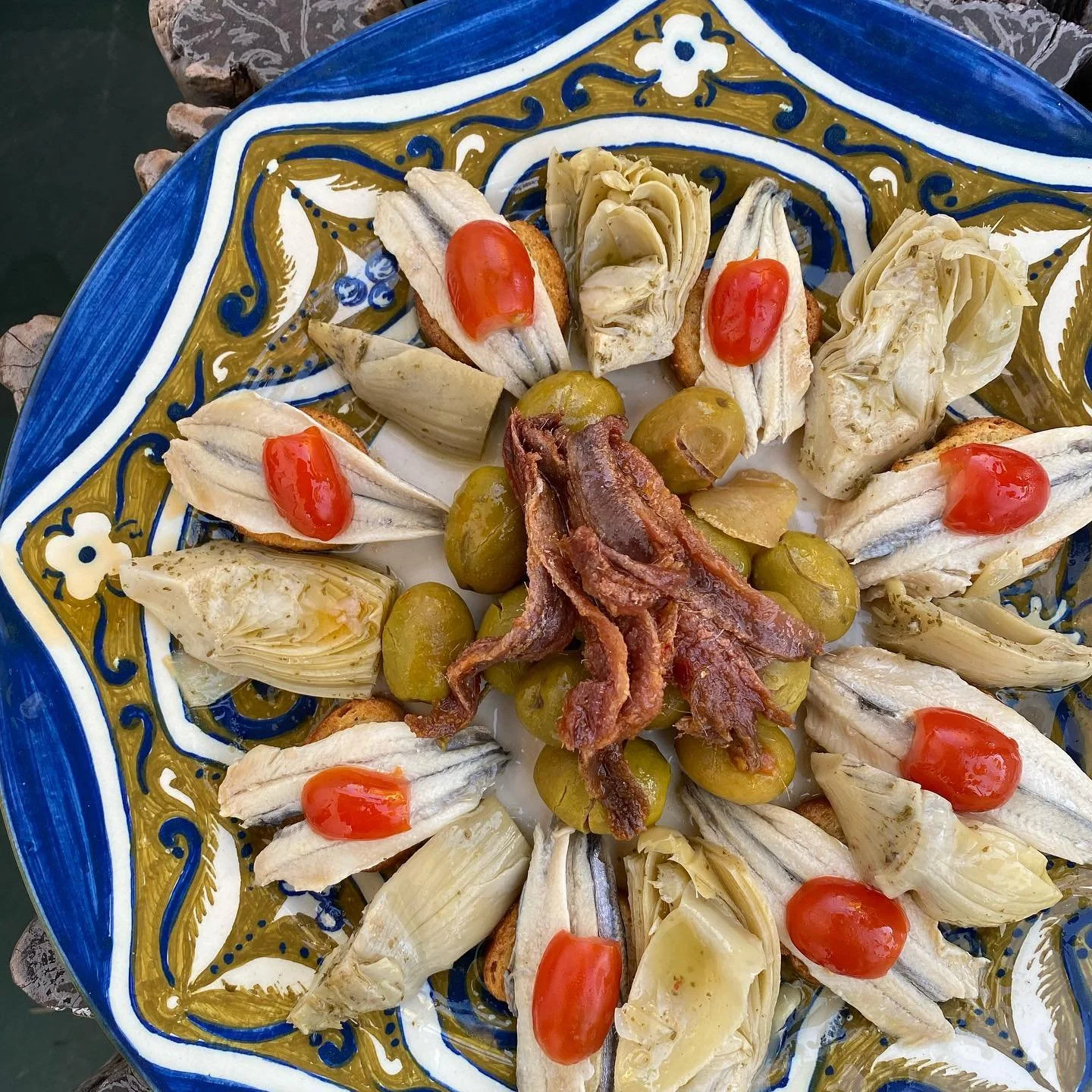 Colored ceramic plate with artful fruit arrangement including peeled bananas topped with cherry tomatoes, artichoke hearts, grapes, and bacon strips in the center, on a decorated surface.