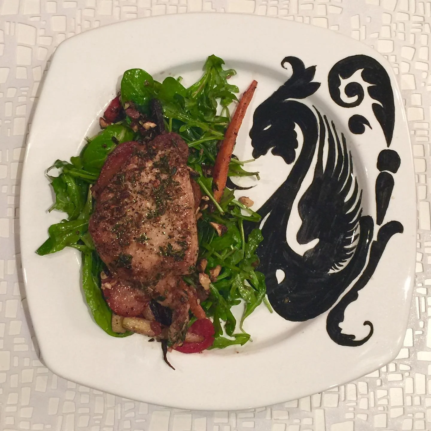 Plate of cooked meat with herbs, served on a bed of mixed greens and vegetables, on a square white plate with a black abstract lion design.