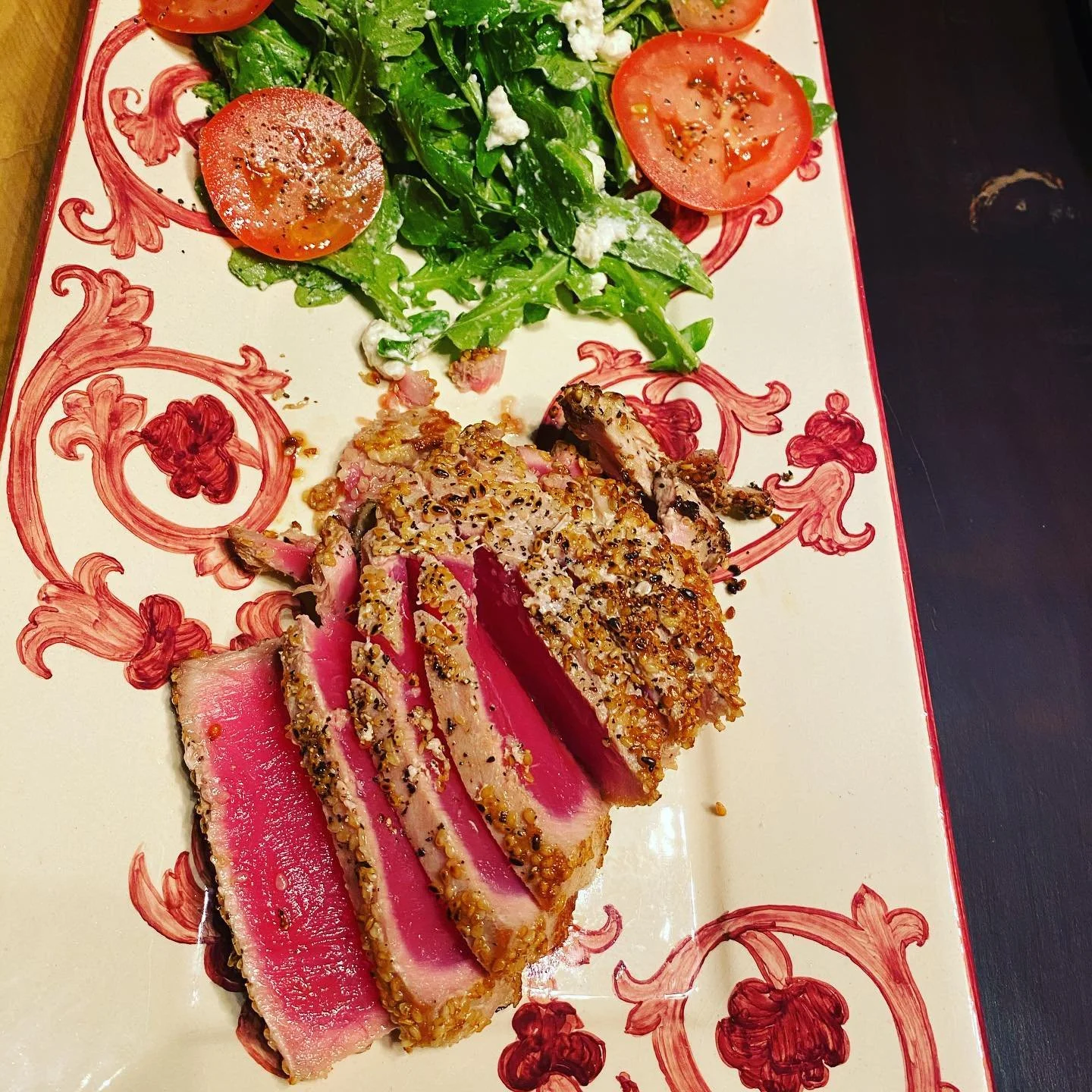 A plate with sliced seared tuna coated in sesame seeds, a fresh green salad with arugula and crumbled feta cheese, and slices of tomato on a decorative rectangular plate.
