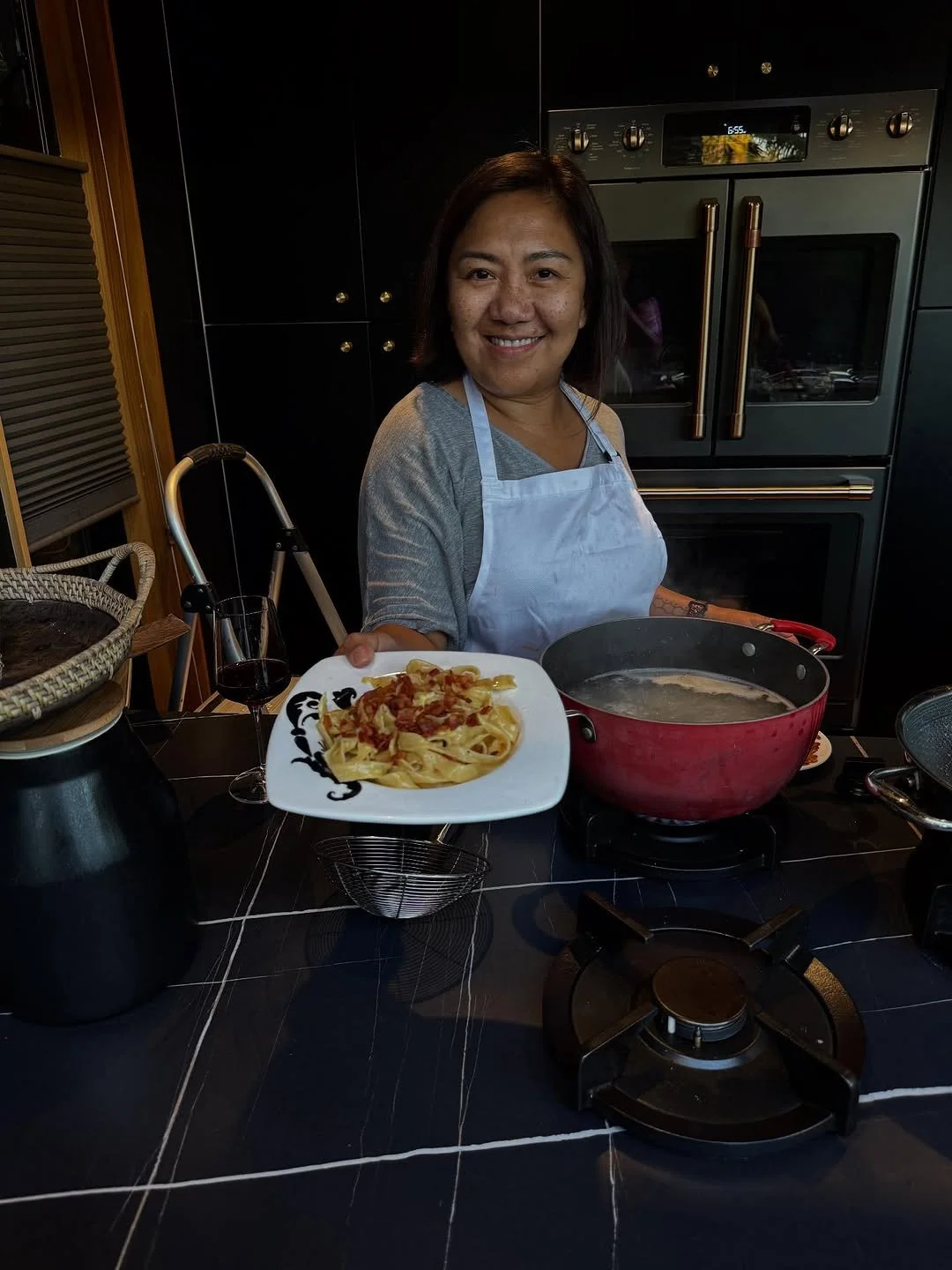 Woman in a kitchen holding a plate of pasta with bacon, smiling, with a glass of red wine nearby.