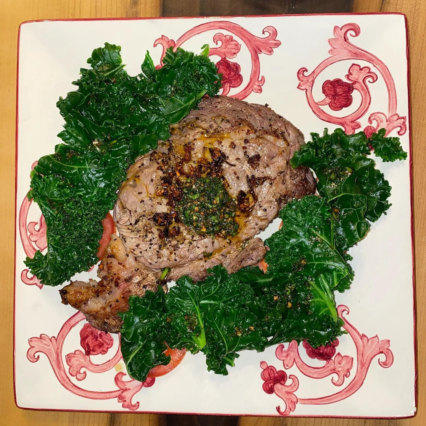 A plate with a cooked beef steak topped with herbs and lights oil, garnished with fresh kale leaves and slices of tomato.