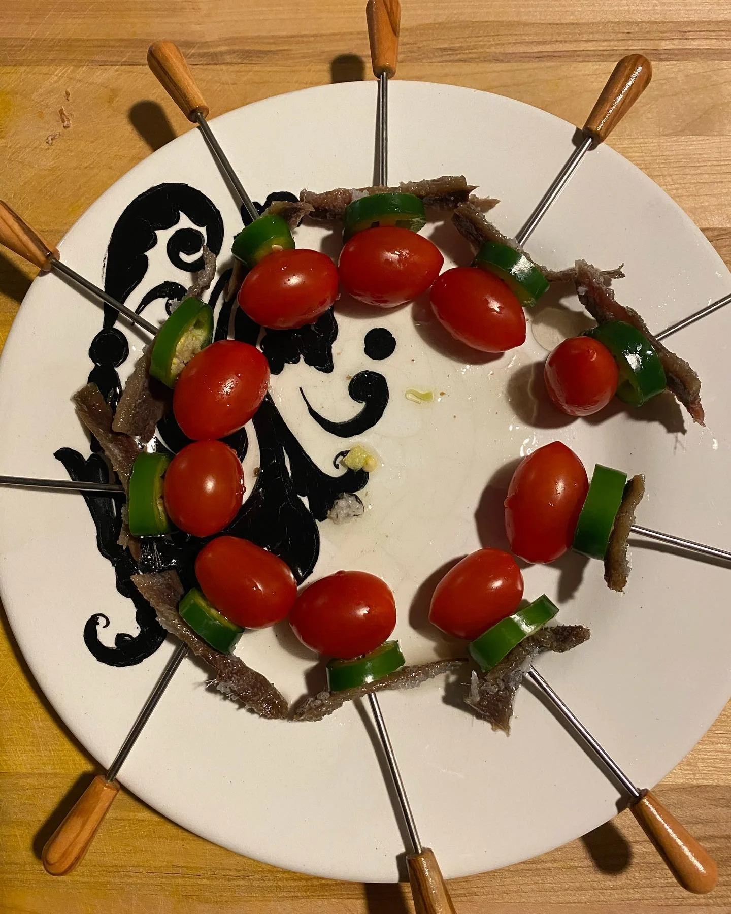 Raw skewers with cherry tomatoes, green peppers, and pieces of dried meat arranged on a plate with a black and white abstract face design.