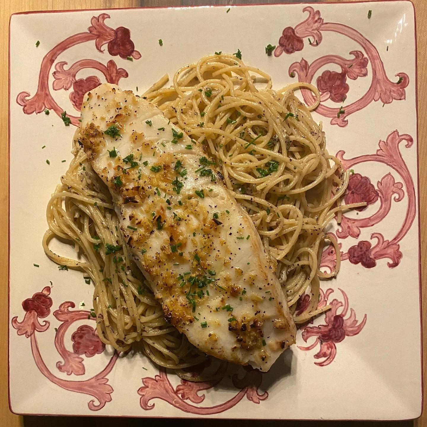 A plate of spaghetti topped with a cooked fish fillet, garnished with chopped herbs and black pepper.