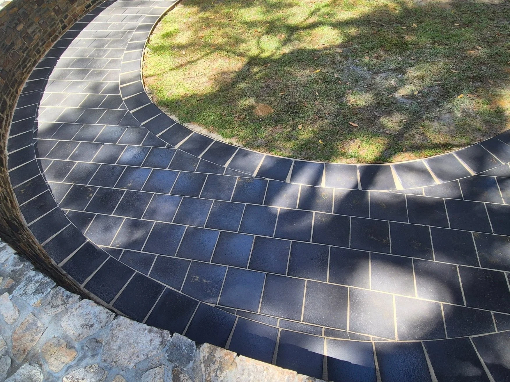 Curved stone pathway with dark rectangular tiles and light grout, surrounding a grassy area with shadows of trees.