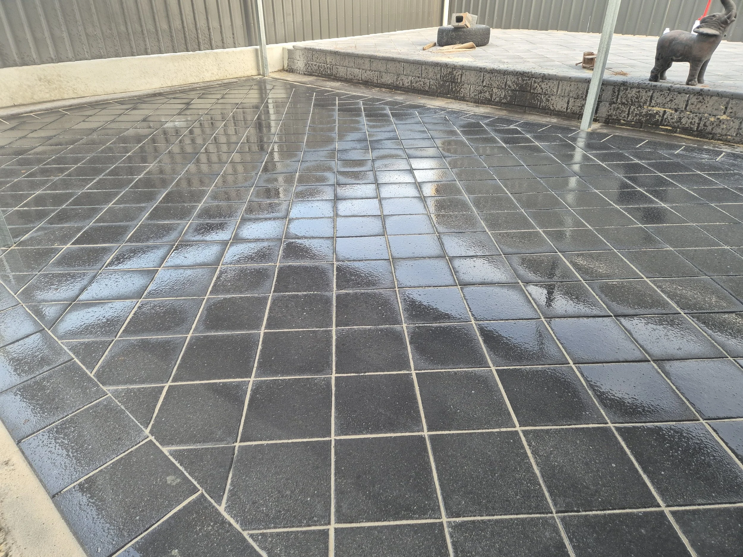 Wet black tiled patio with reflections of the sky, fence, and objects, including a tire, on the surface.