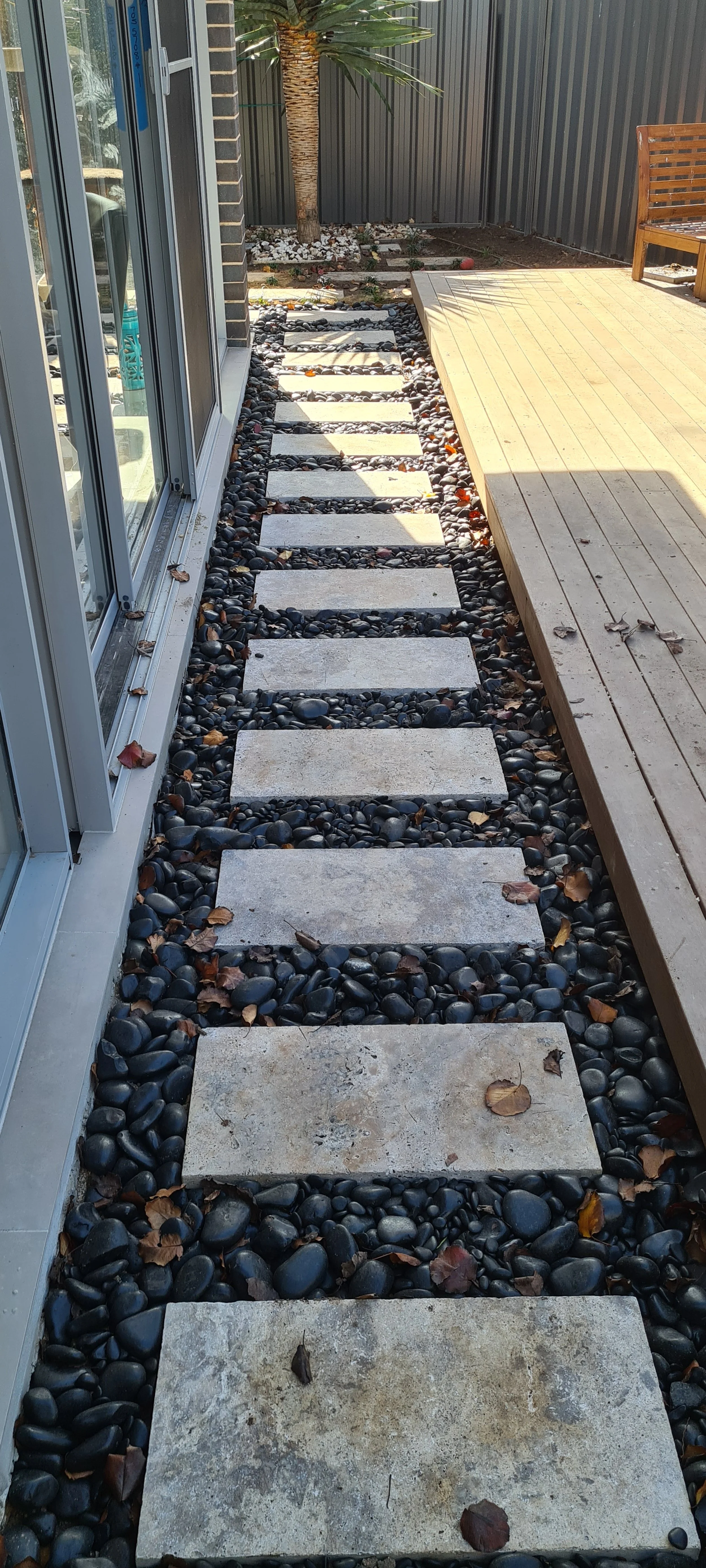 A backyard with a sliding glass door, small step paving stones on black pebbles, a wooden deck, a sitting bench, and a small tree with mulch.