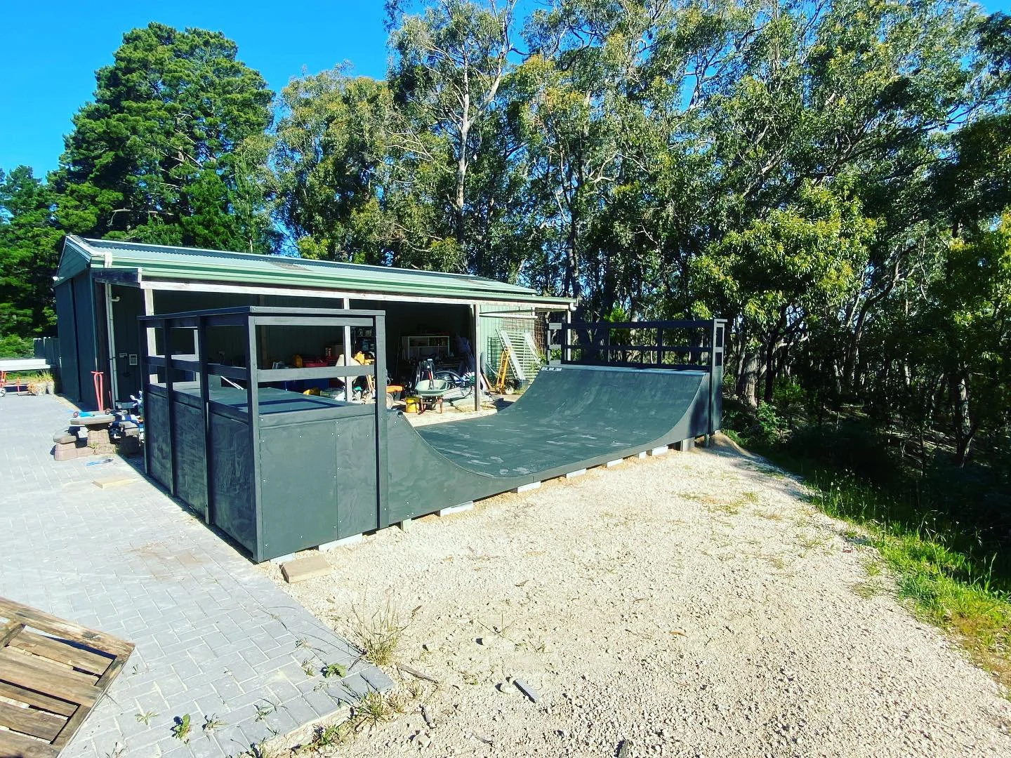 A backyard with a black skate ramp next to a shed, surrounded by green trees on a sunny day.