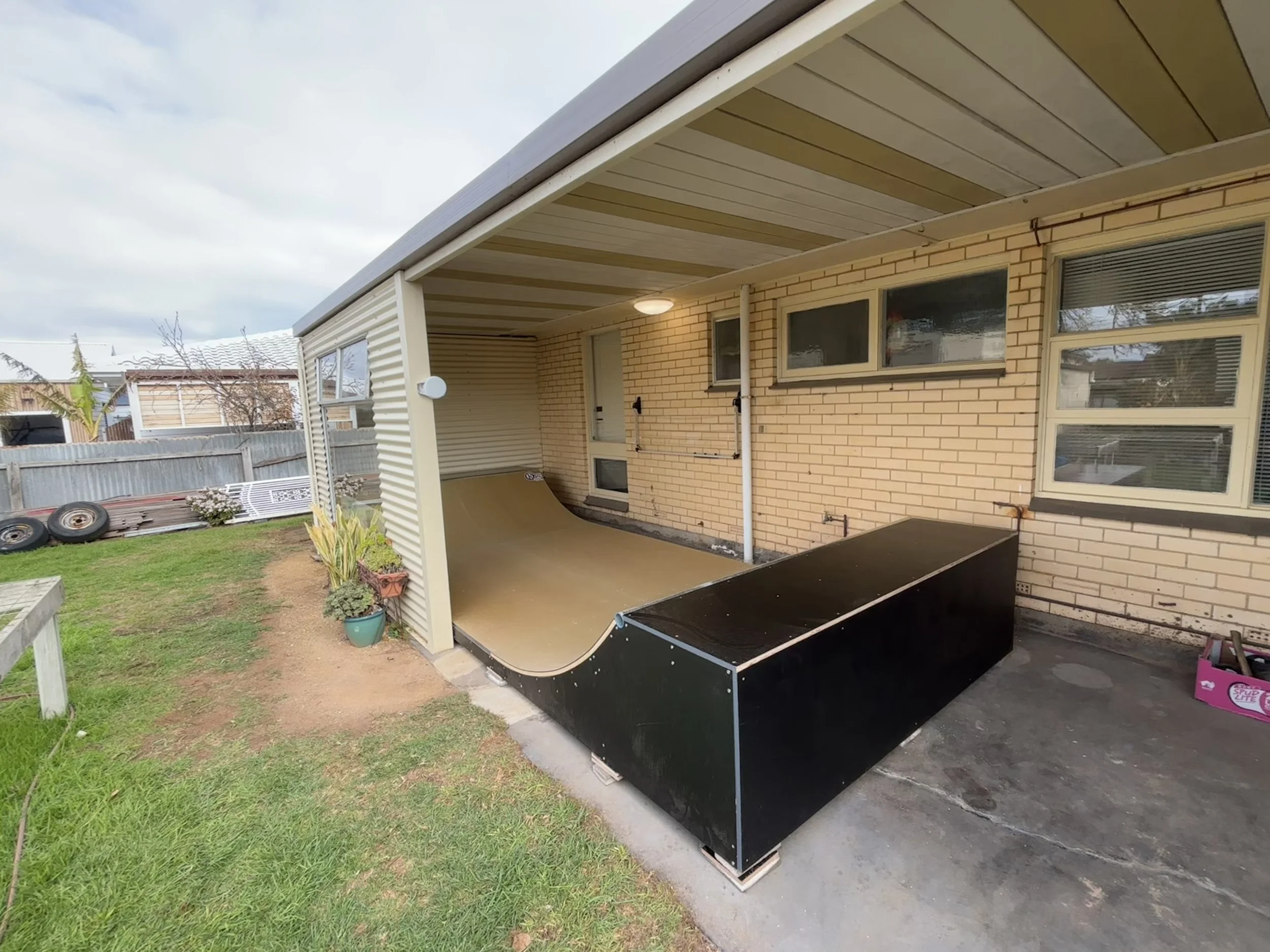 Backyard patio with a mini skate ramp, potted plants, and tires against a wooden fence.