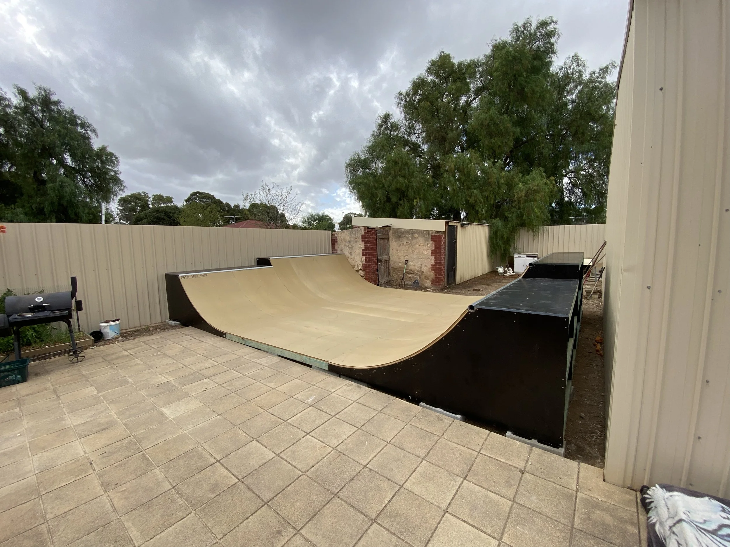A backyard skate ramp constructed with a wooden surface, black sides, on a paved patio area, enclosed by a beige fence, with trees, a cloudy sky, and a shed with a brick wall in the background.