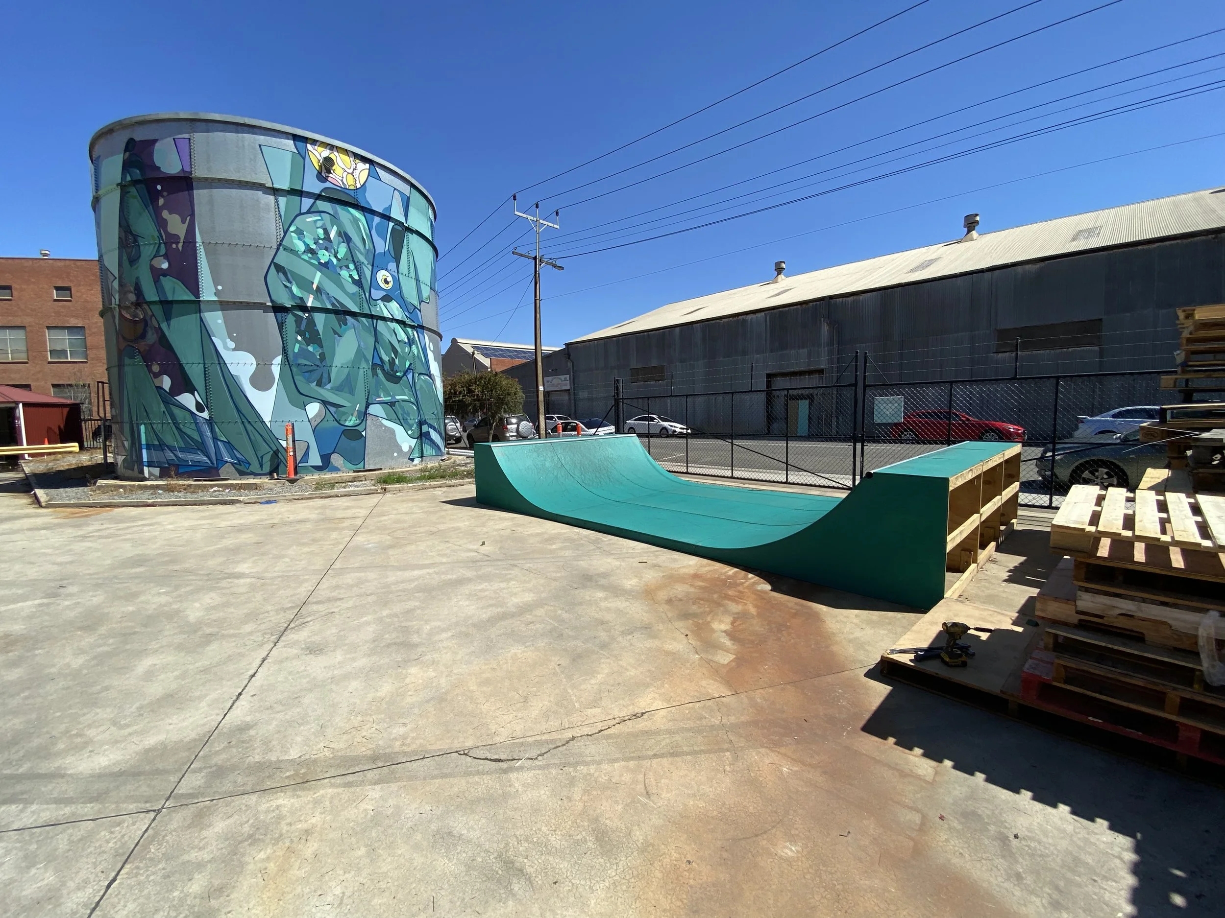 Outdoor skatepark with a teal half-pipe ramp, adjacent to a colorful cylindrical building with a mural of a green creature, and a clear blue sky in the background.