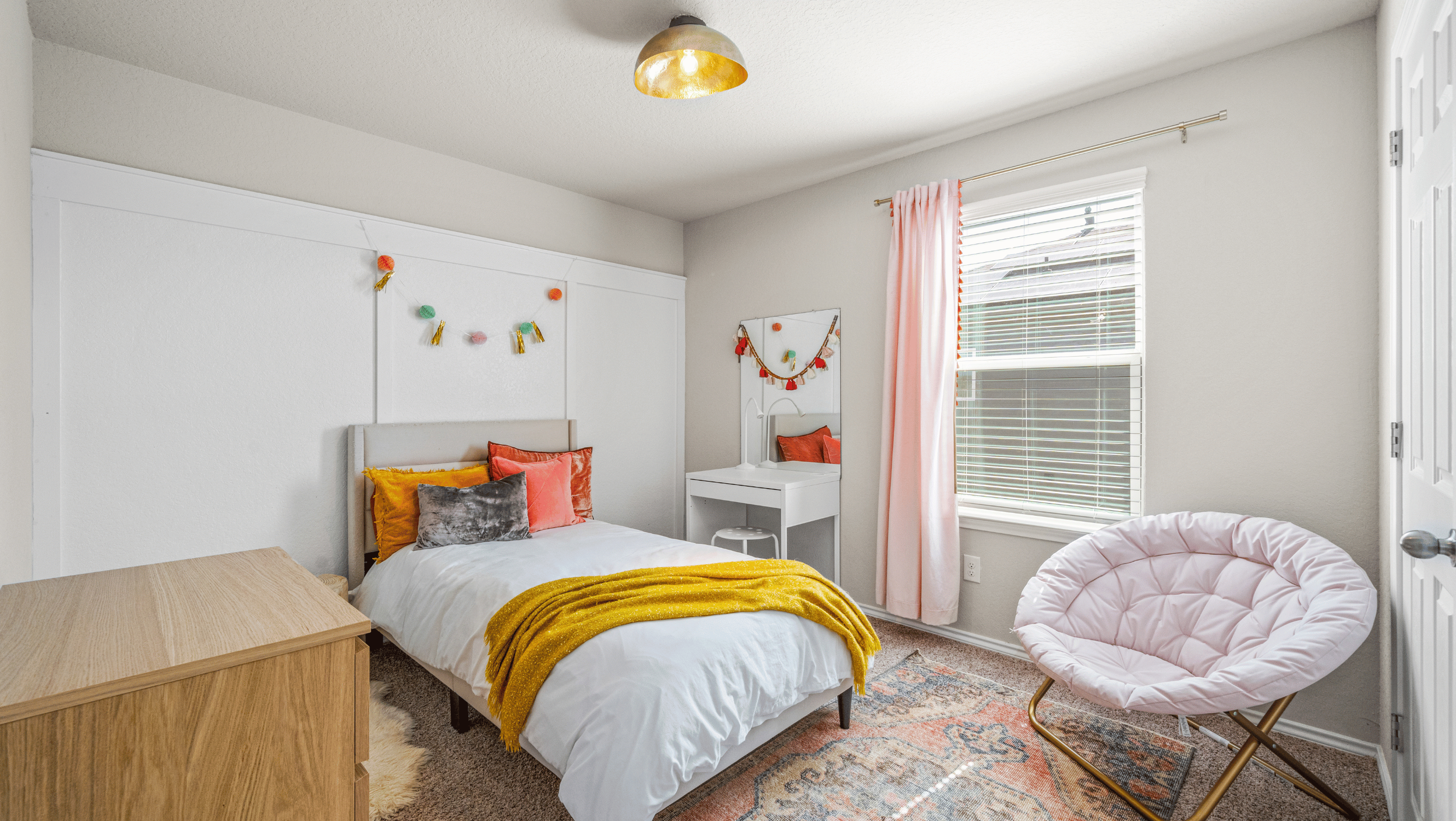 Cozy bedroom with a single bed, colorful pillows, and a yellow throw. A white vanity set with a mirror and a stool is against the wall. A pink papasan chair and a wood dresser are in the room. Decorative garlands hang on the wall and mirror. Light pi