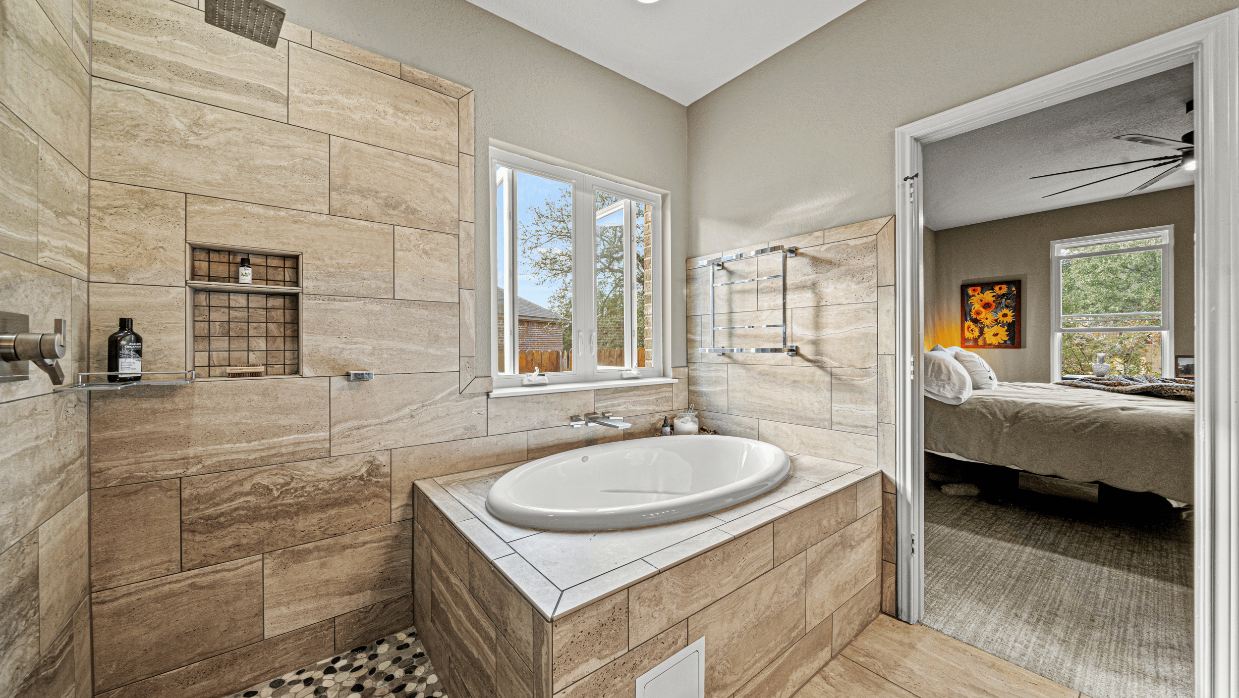 Modern bathroom with a large bathtub, stone tile walls, a window, and a view into a bedroom with a bed and sunflower artwork.
