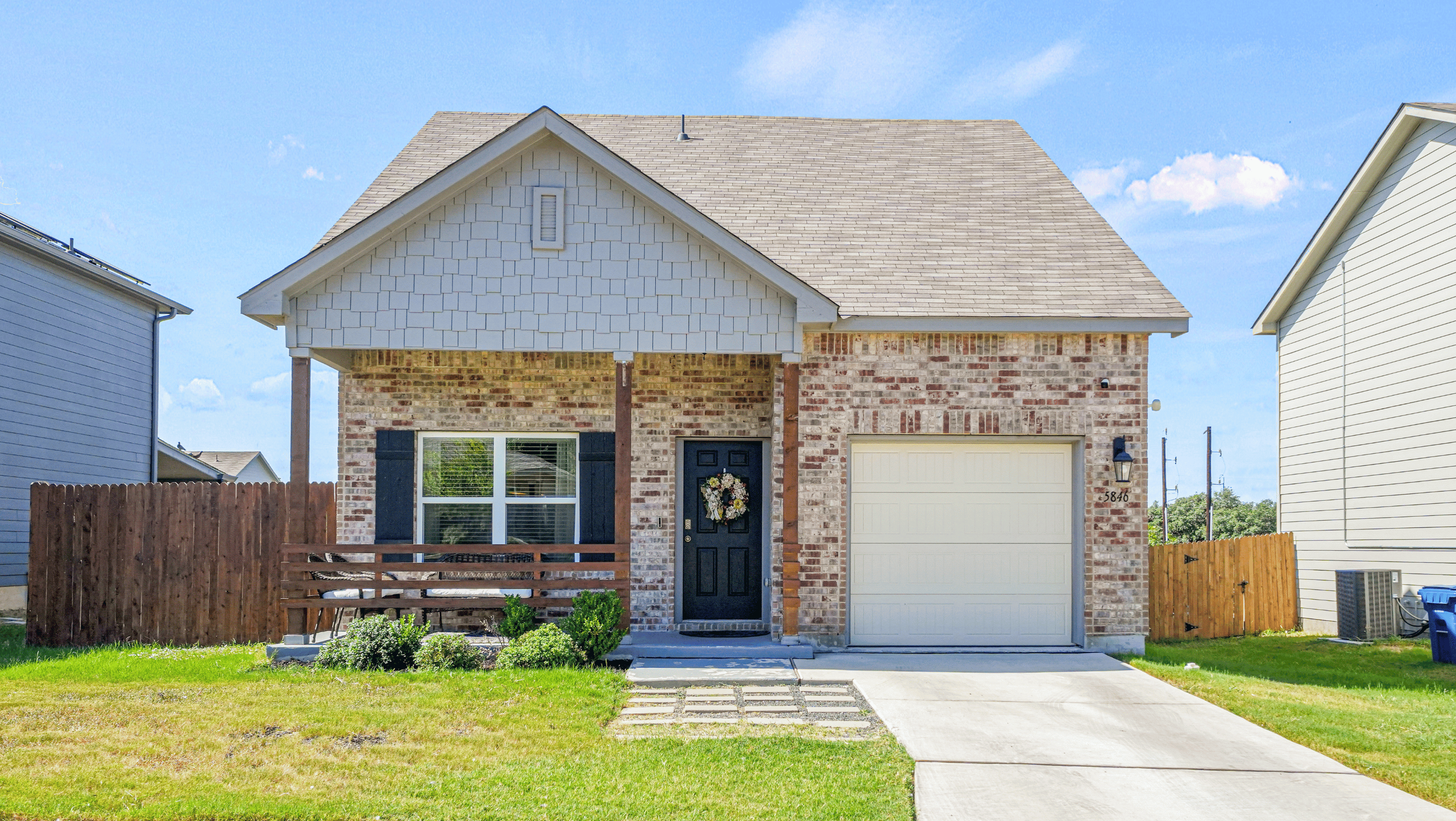 Single-story brick house with a covered porch, attached garage, and front yard with green grass. The entrance features a black door with a decorative wreath.