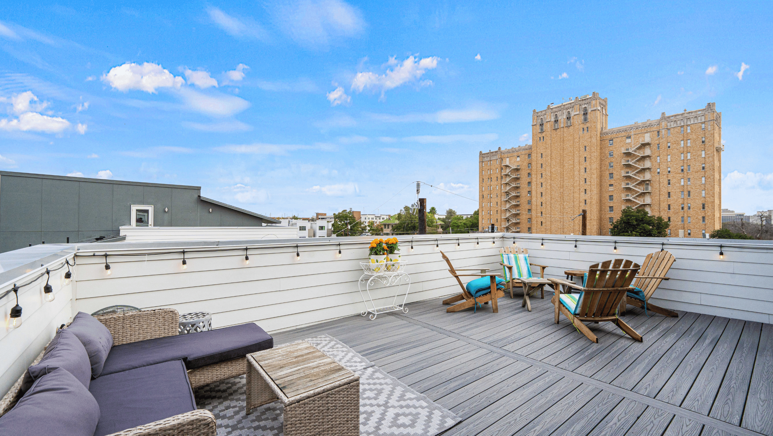 Rooftop patio with wicker furniture, cushions, and a wooden table. String lights hang along white railing. A potted plant with yellow flowers is on a white stand. A tall brick building is visible in the background.