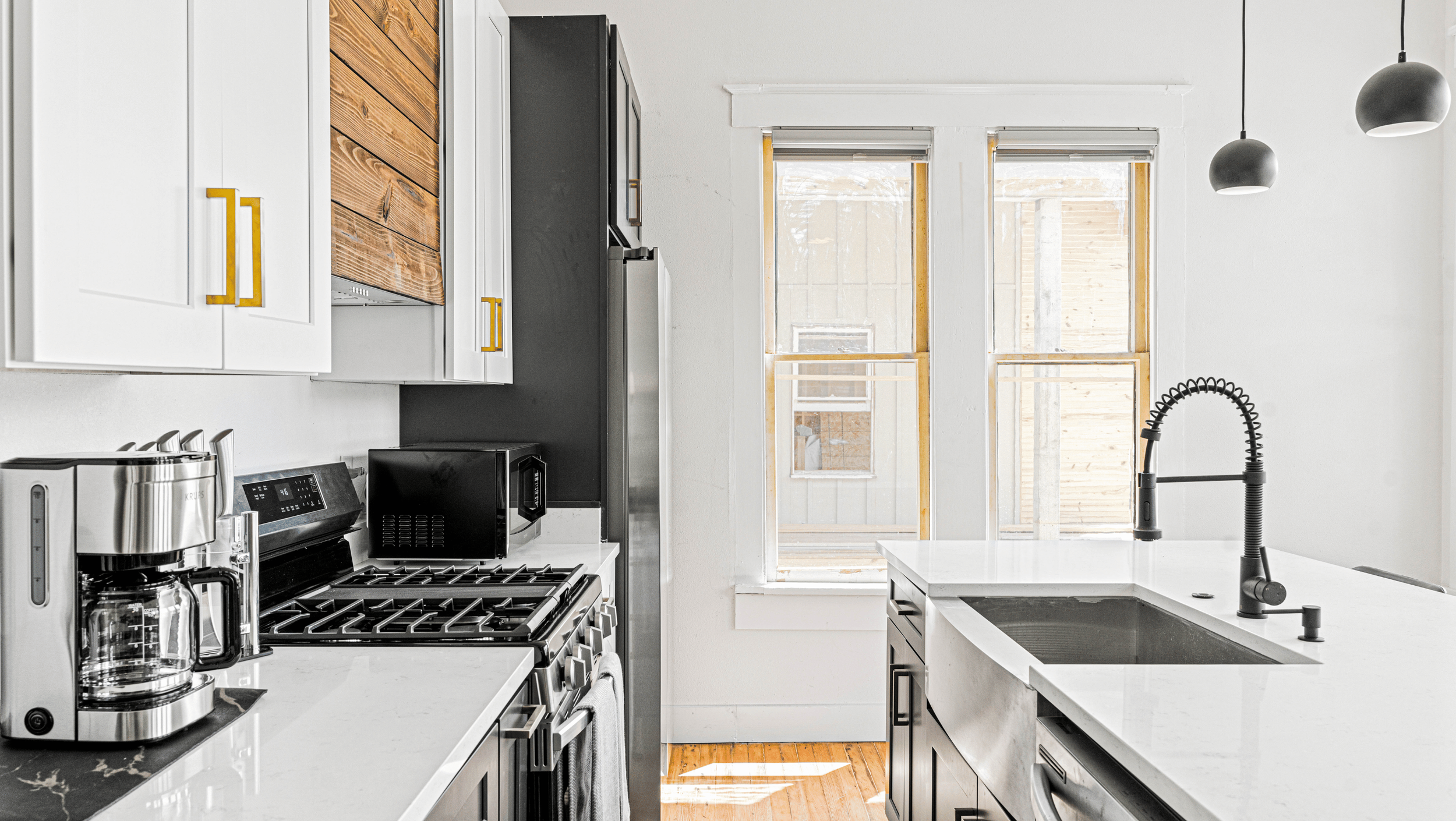 Modern kitchen with white cabinets, stainless steel appliances, and black accents, featuring a coffee maker, stove, microwave, and sleek sink with a spiral faucet.