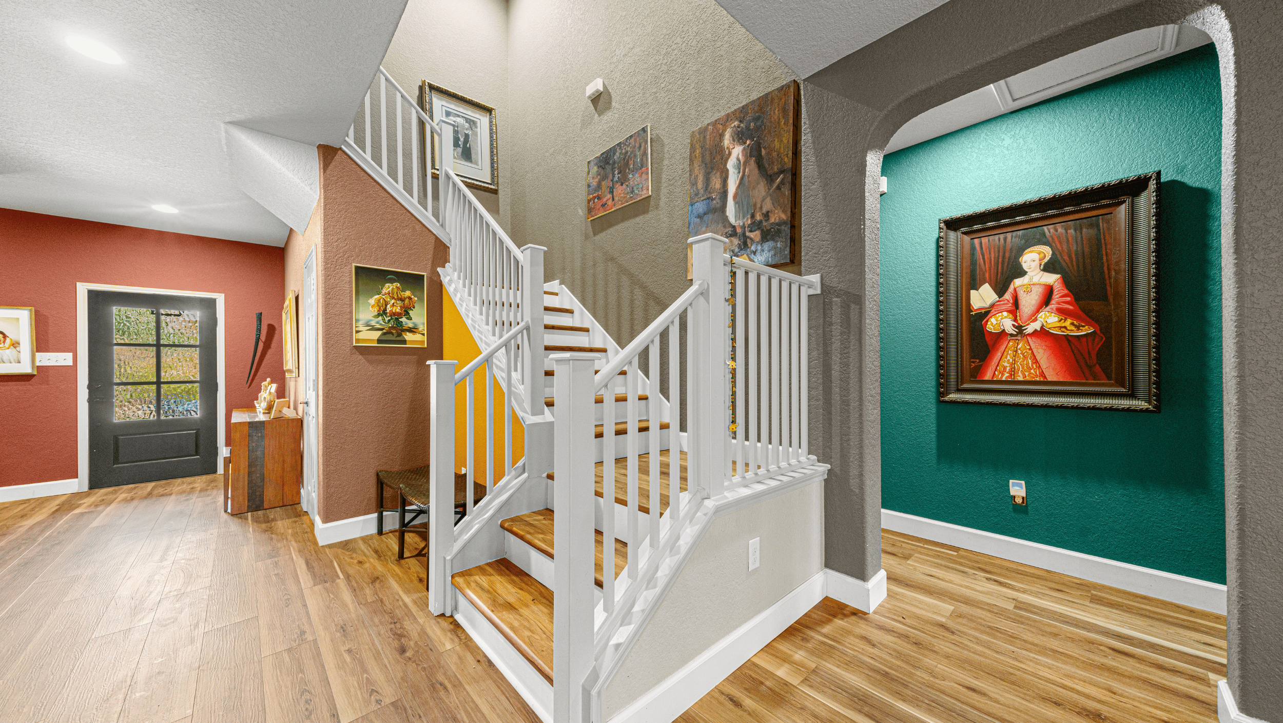 A modern interior with a wooden staircase and colorful walls featuring various paintings. The staircase has white railings, and the floor is wood. A green wall displays a framed painting of a woman in red. The space is well-lit with recessed lighting