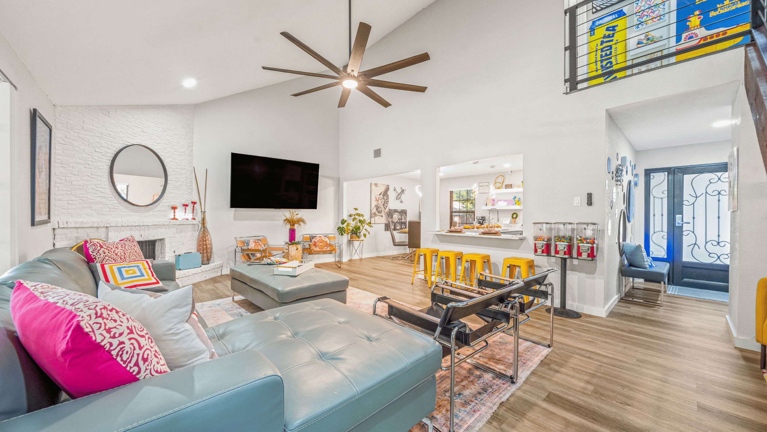 Modern living room with vaulted ceiling, blue sofa, colorful throw pillows, a large wall-mounted TV, and a bar area with yellow stools. White brick fireplace with a round mirror, contemporary decor, and wooden floors. Bright interior with eclectic de