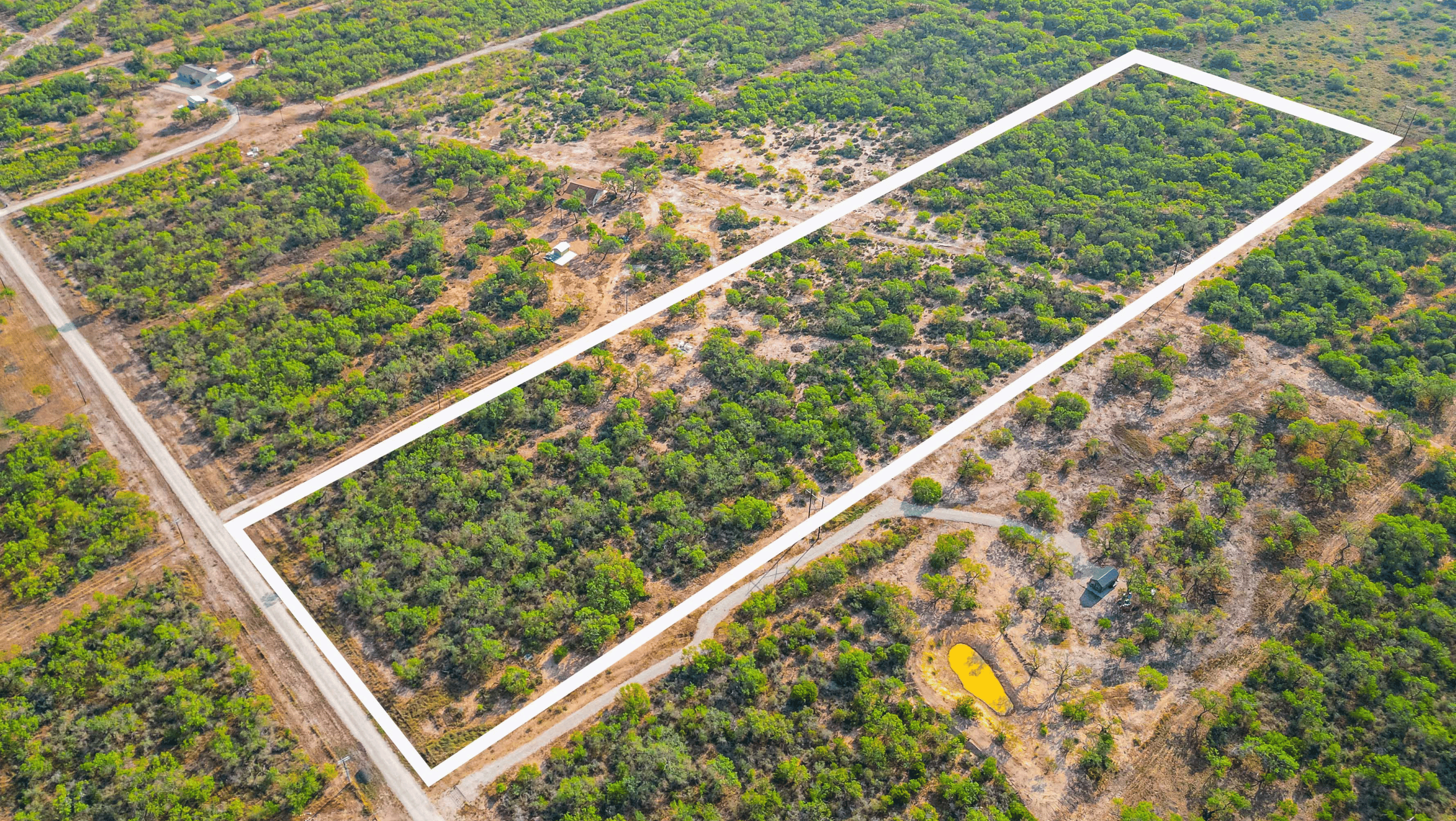 Aerial view of a rectangular plot of land marked in white, surrounded by trees and vegetation. Nearby are smaller structures and paths visible in the landscape.