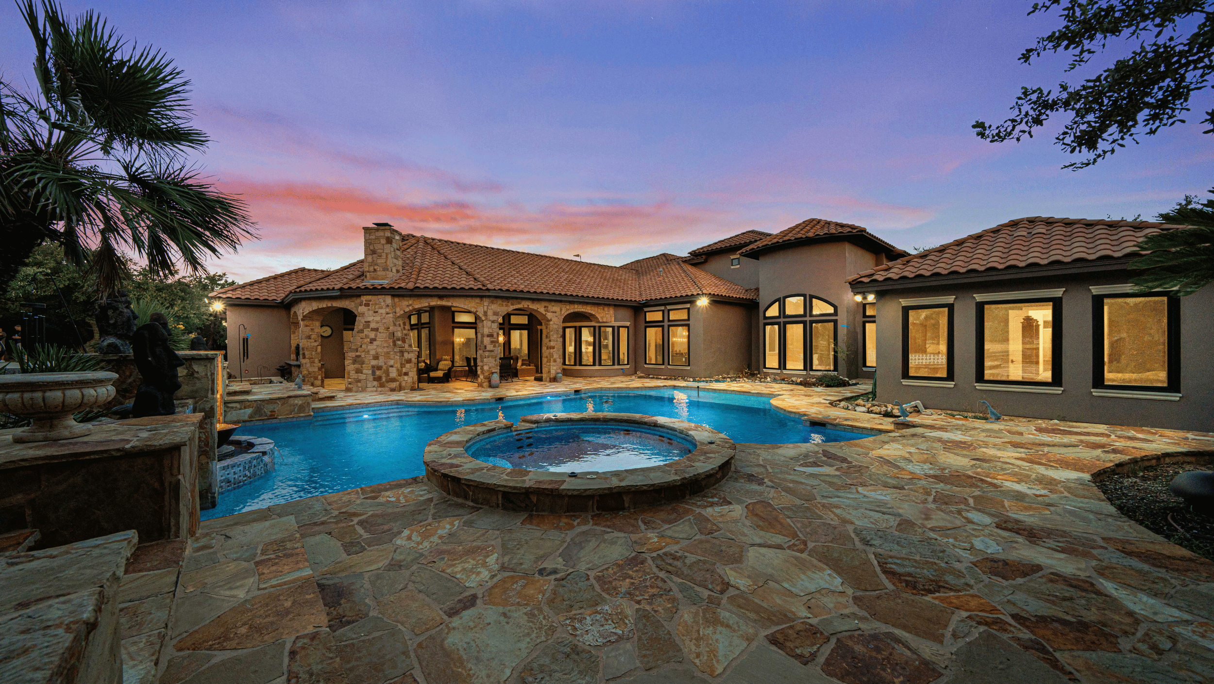 Luxurious home with swimming pool and spa in backyard at dusk, featuring Mediterranean-style architecture with stone tiles and terracotta roof.