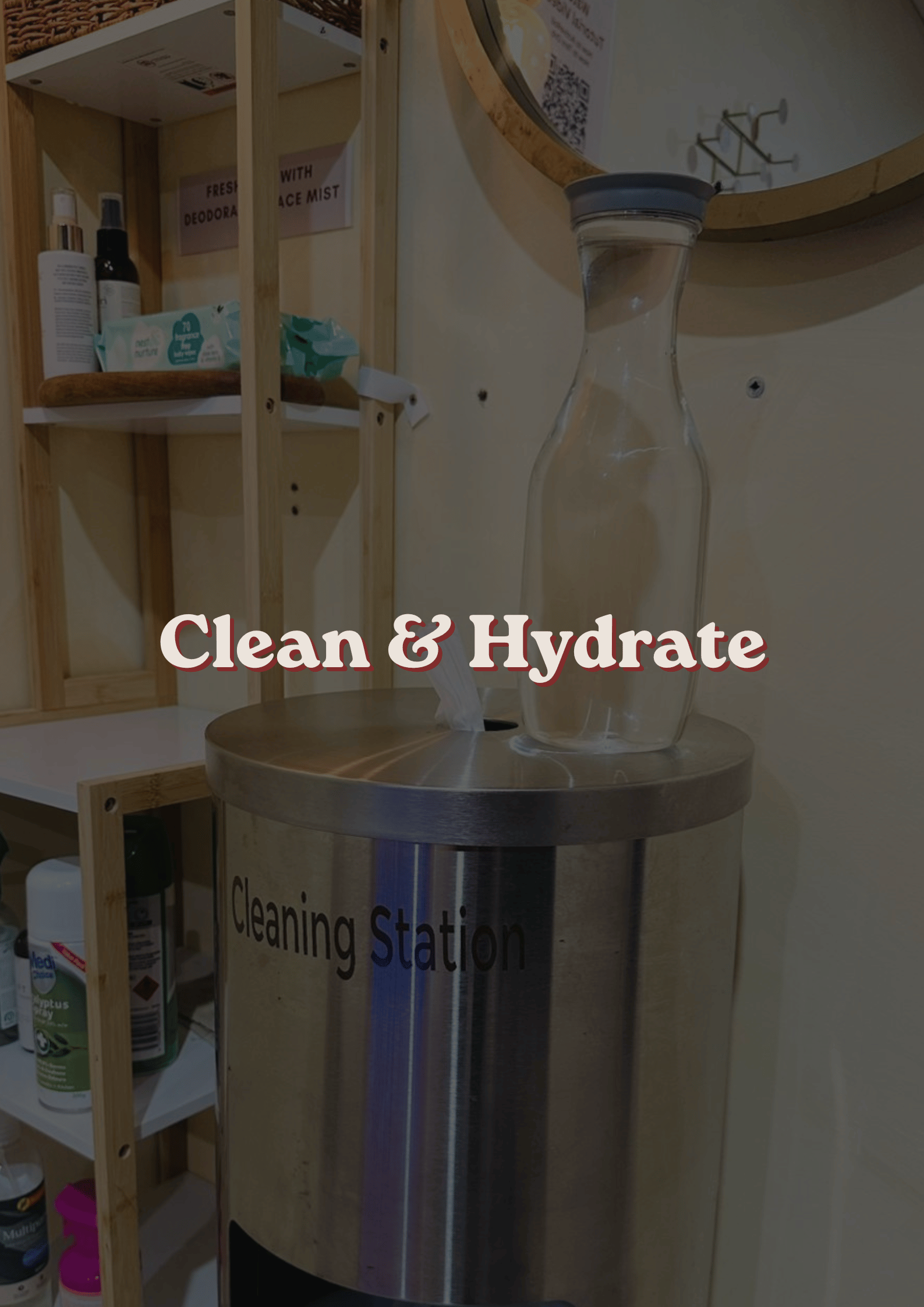 A transparent tissue box and a stainless steel contactless hand sanitizer or cleaning station with a deep round top, situated on a wooden counter in a store or office space. Wooden shelving with various hygiene products and signs are visible in the background.