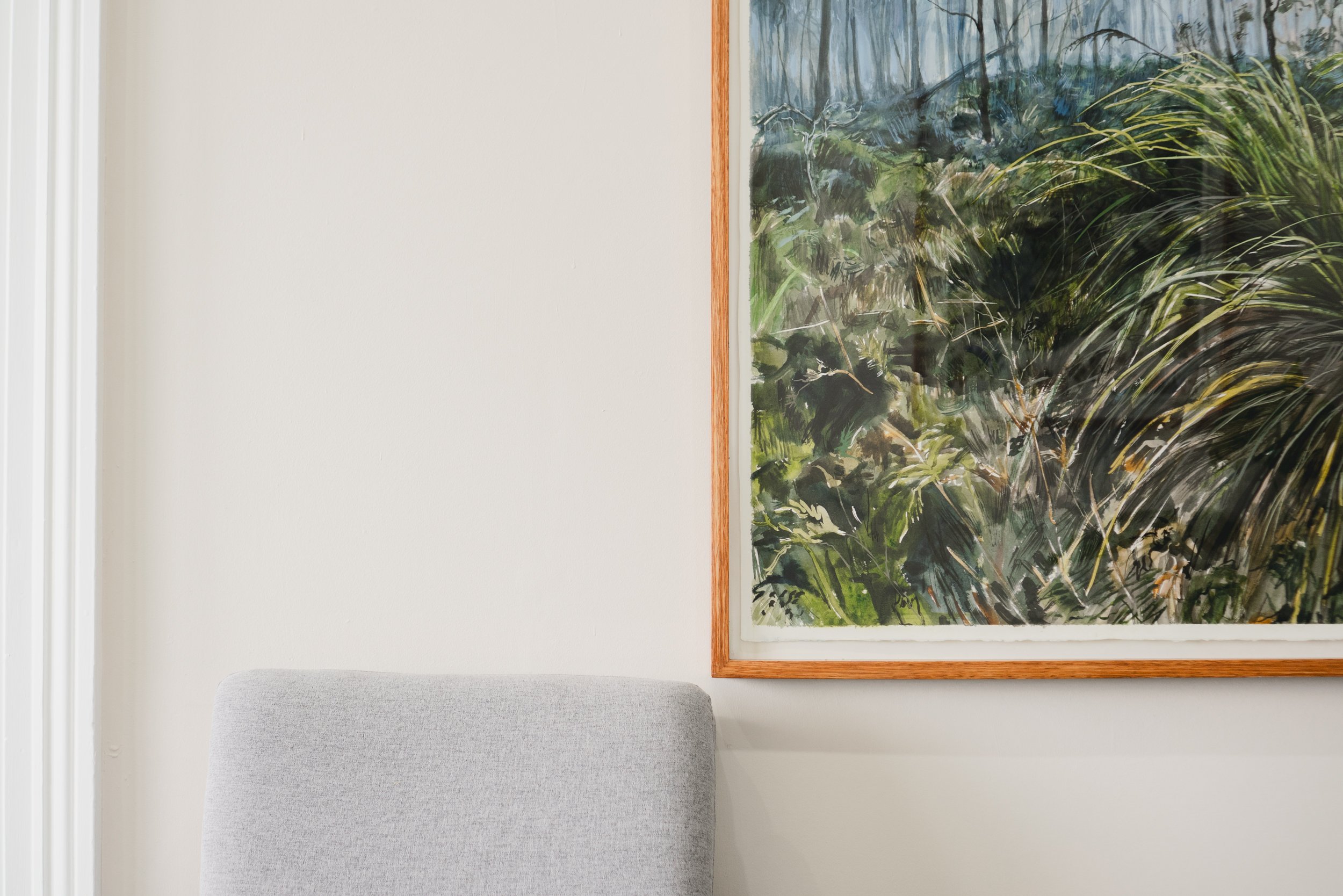 Interior wall with a framed painting of a forest scene and part of a gray upholstered chair.