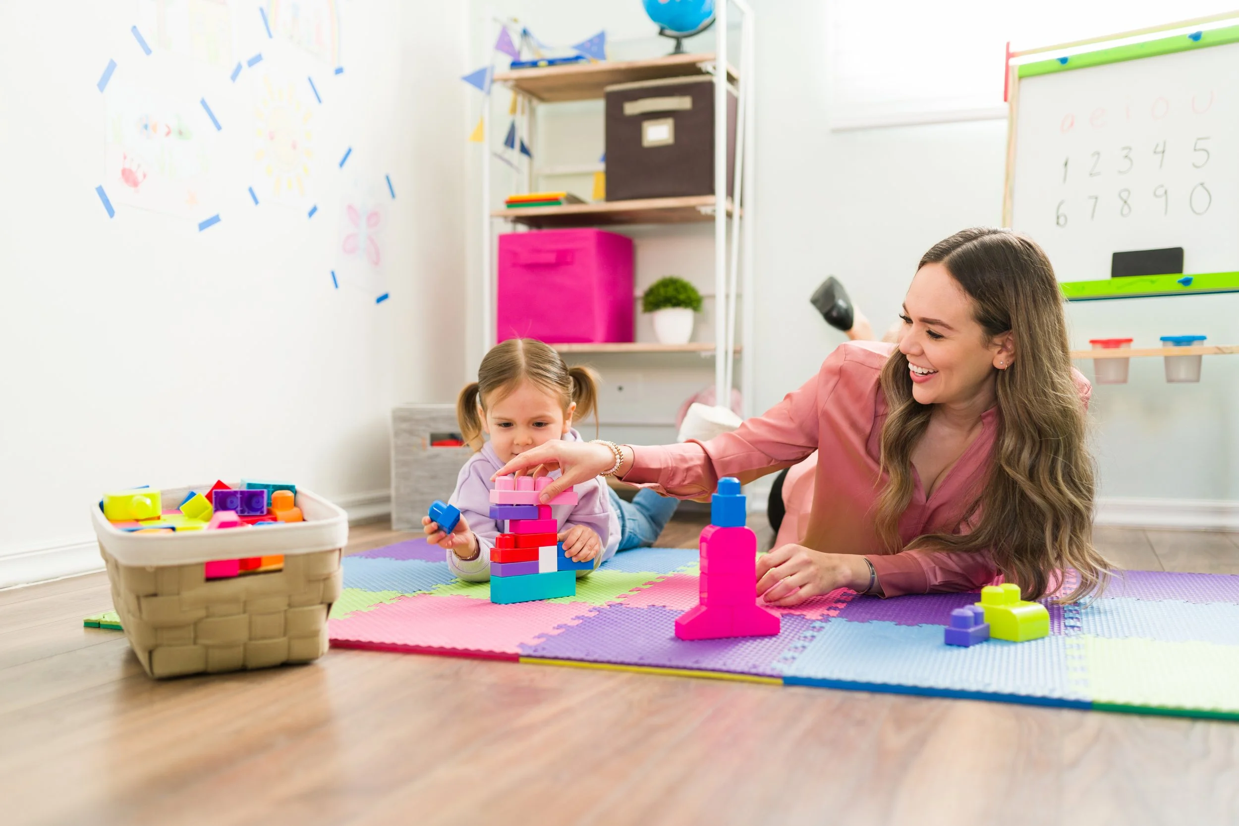 A woman and a young girl playing with colorful building blocks on a foam mat in a playroom.
