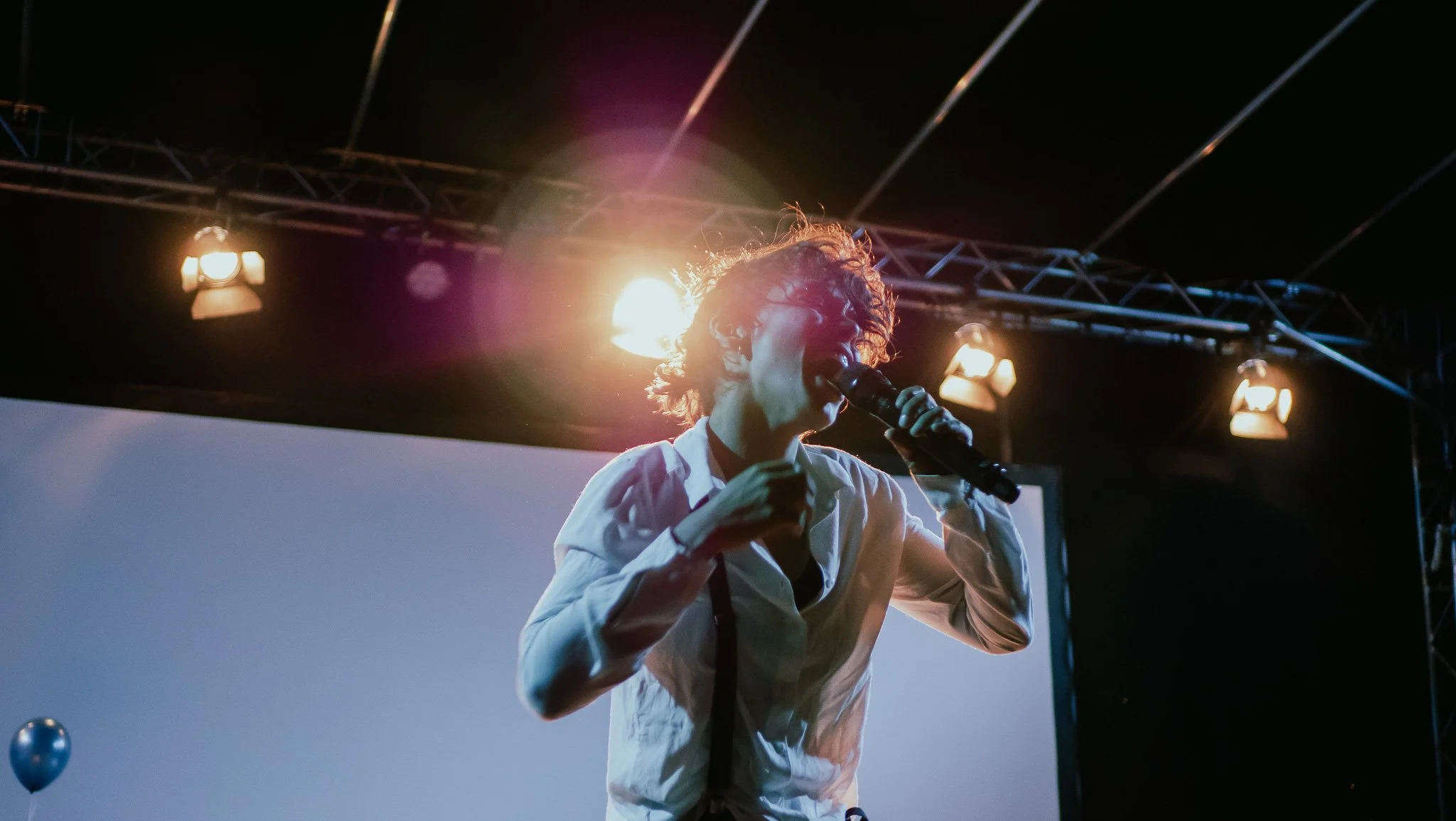 A person with curly hair singing into a microphone on stage with bright stage lights behind them.