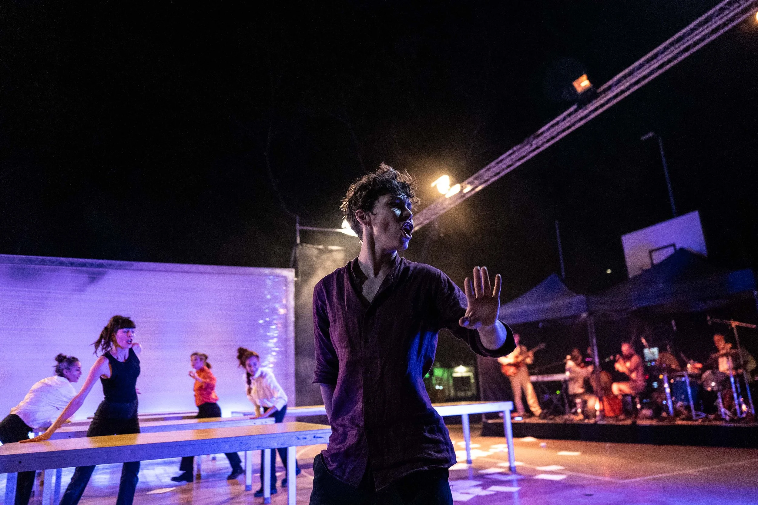Performer in dark shirt gesturing on stage at nighttime with dancers and band in the background.