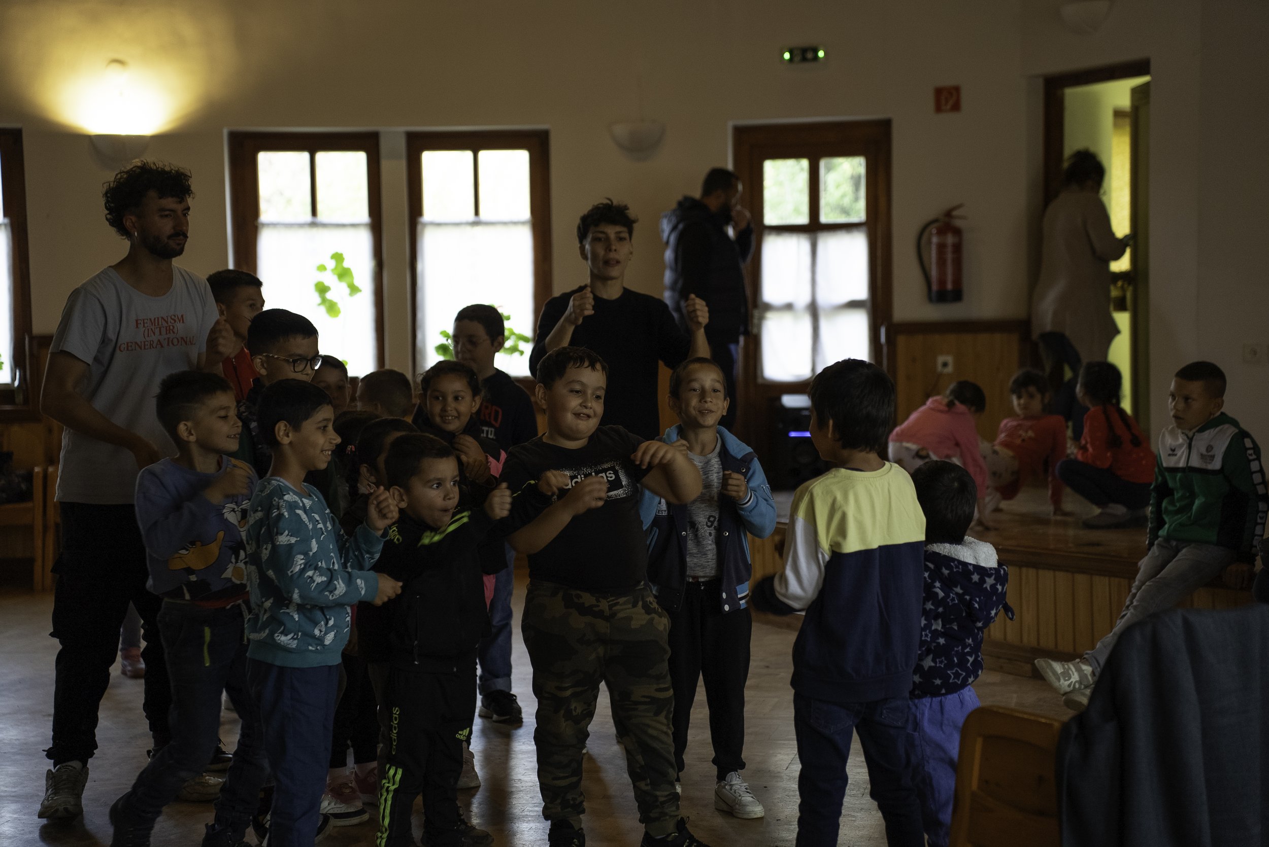 Group of children and two adults in a playroom or community center, with some children standing and others sitting on a stage, engaging in activities or games, with wooden floors and natural light from windows.