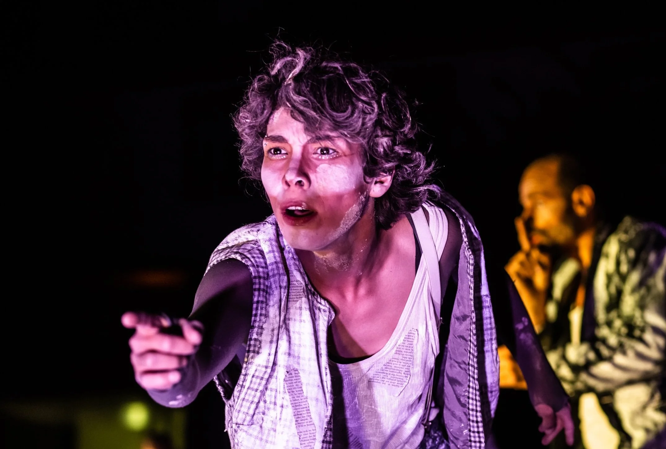 A woman with curly hair, expressive face, and vibrant makeup, pointing forward during a stage performance. A man in the background with a finger on his lips, partially visible.