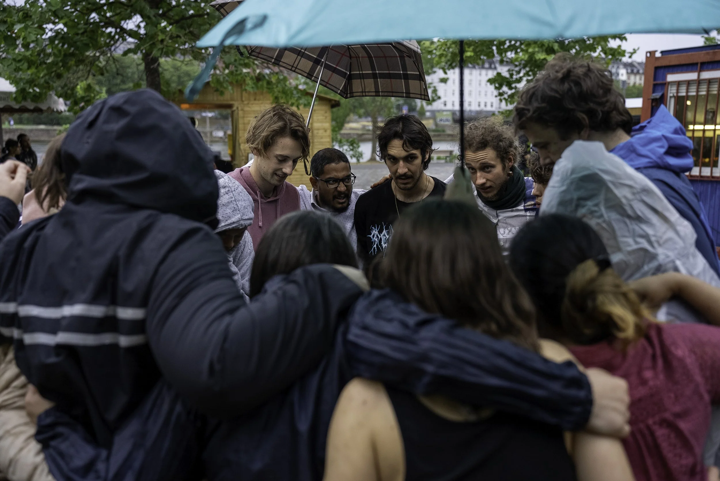 A group of people huddled together outdoors under an umbrella, appearing to be engaged in an activity or discussion.