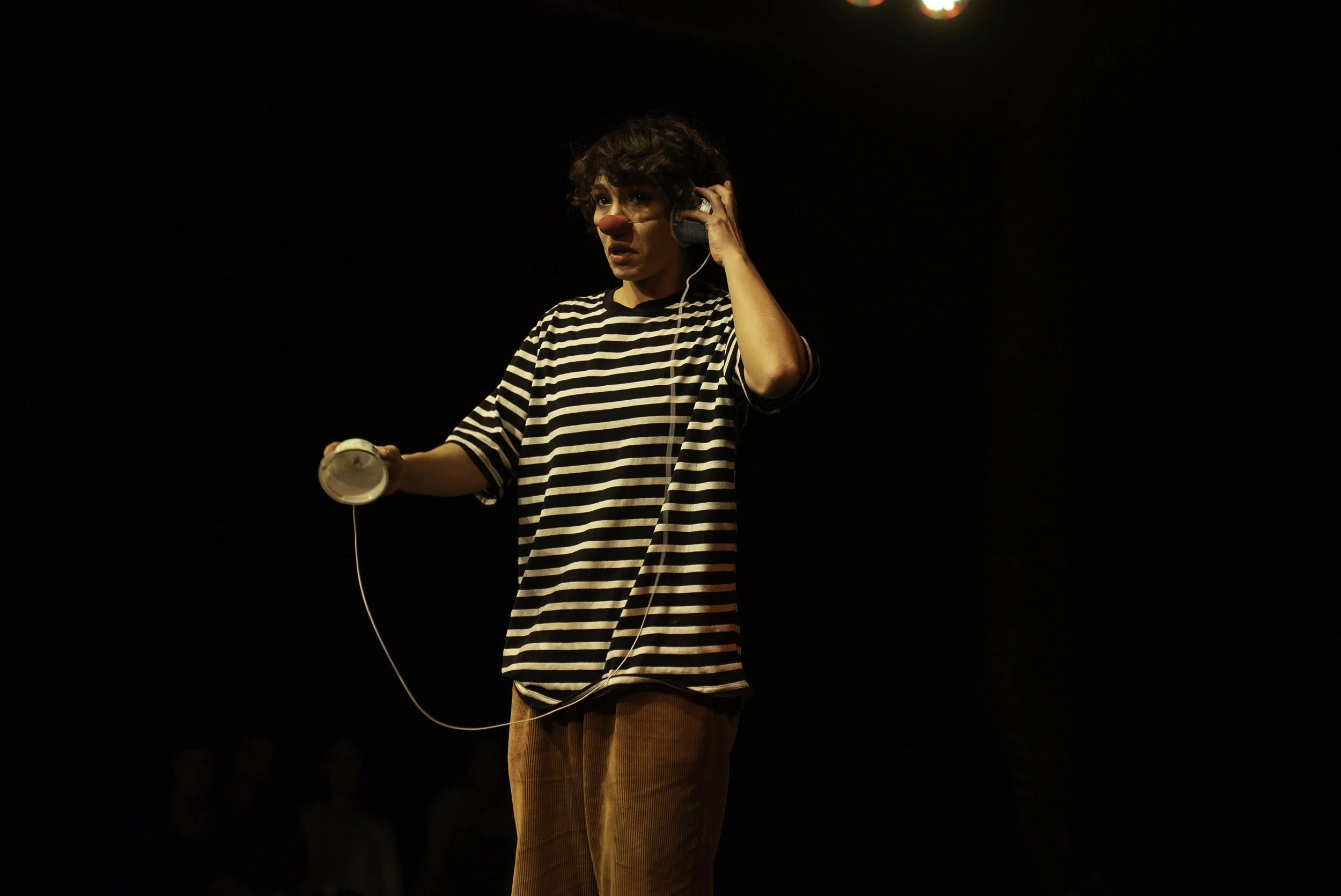 A performer dressed as a clown with a red nose is on stage, wearing a striped shirt and brown pants, holding a makeup container in one hand and wearing headphones, with a dark background and audience in the shadows.
