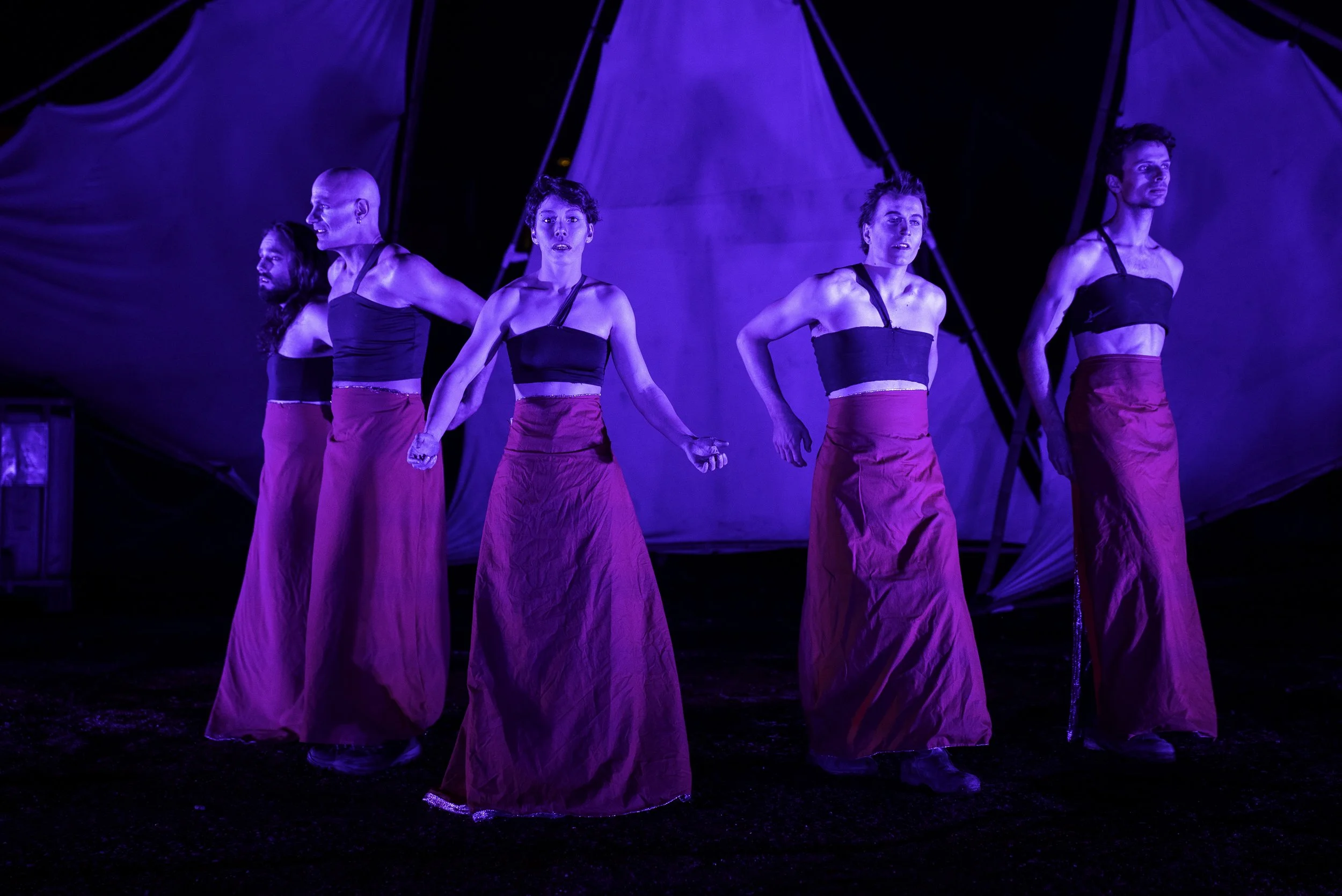 Five performers in black tops and long red skirts holding hands on stage with blue lighting and a tent-like backdrop.