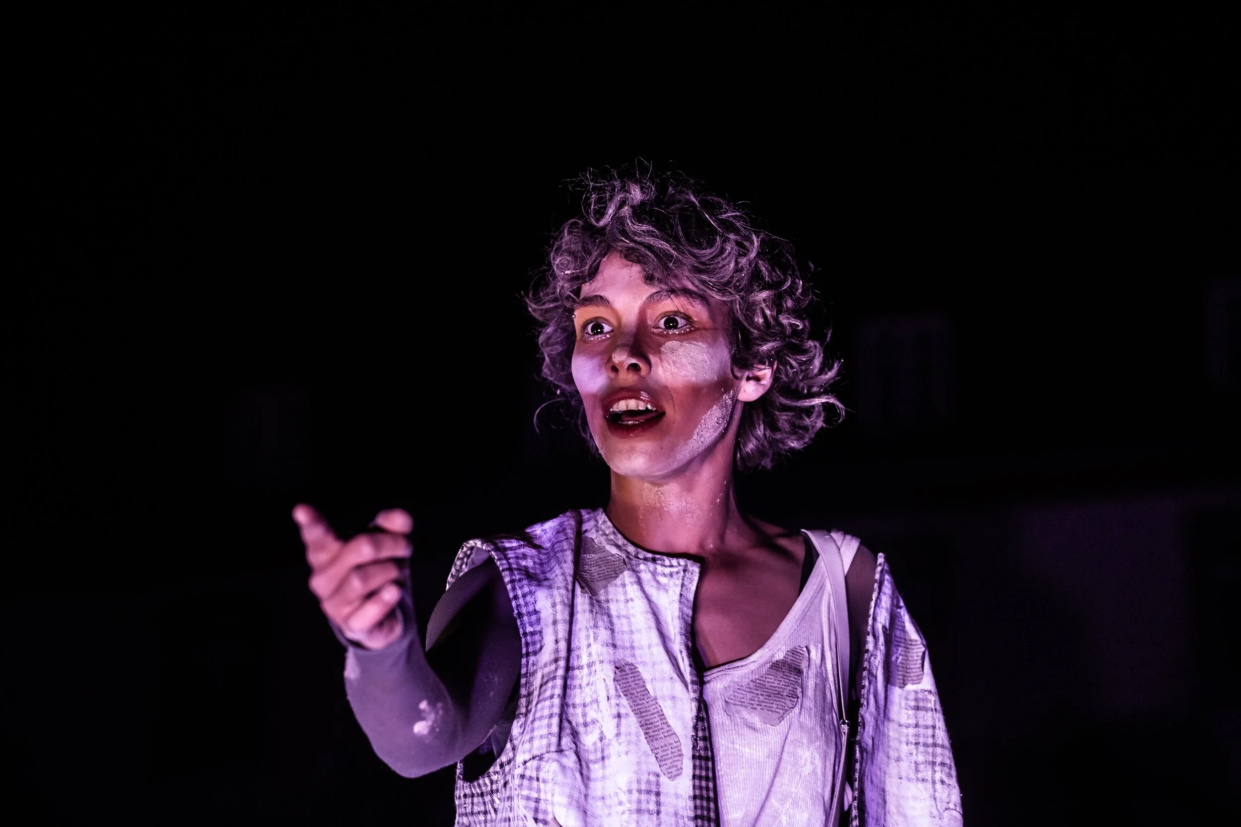 A woman with curly gray hair and face paint looking surprised or intense, pointing forward in a dark setting.