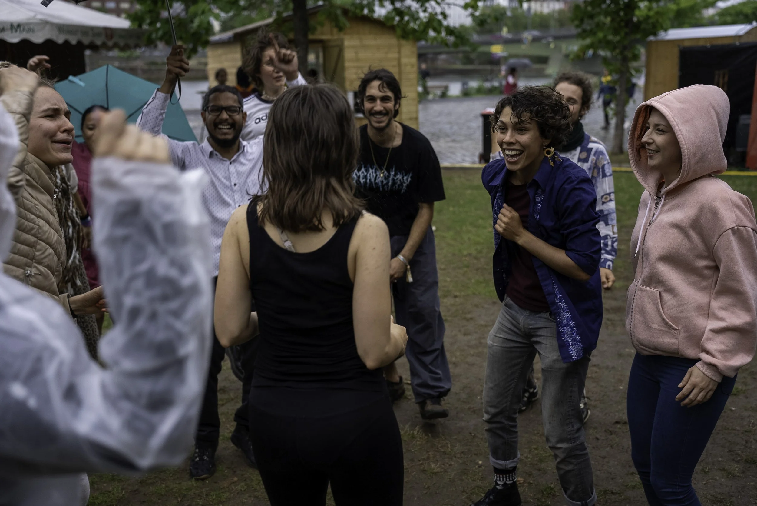 Group of people outdoors, smiling and laughing, engaging in a lively conversation or dance, with trees, grass, and tents in the background.
