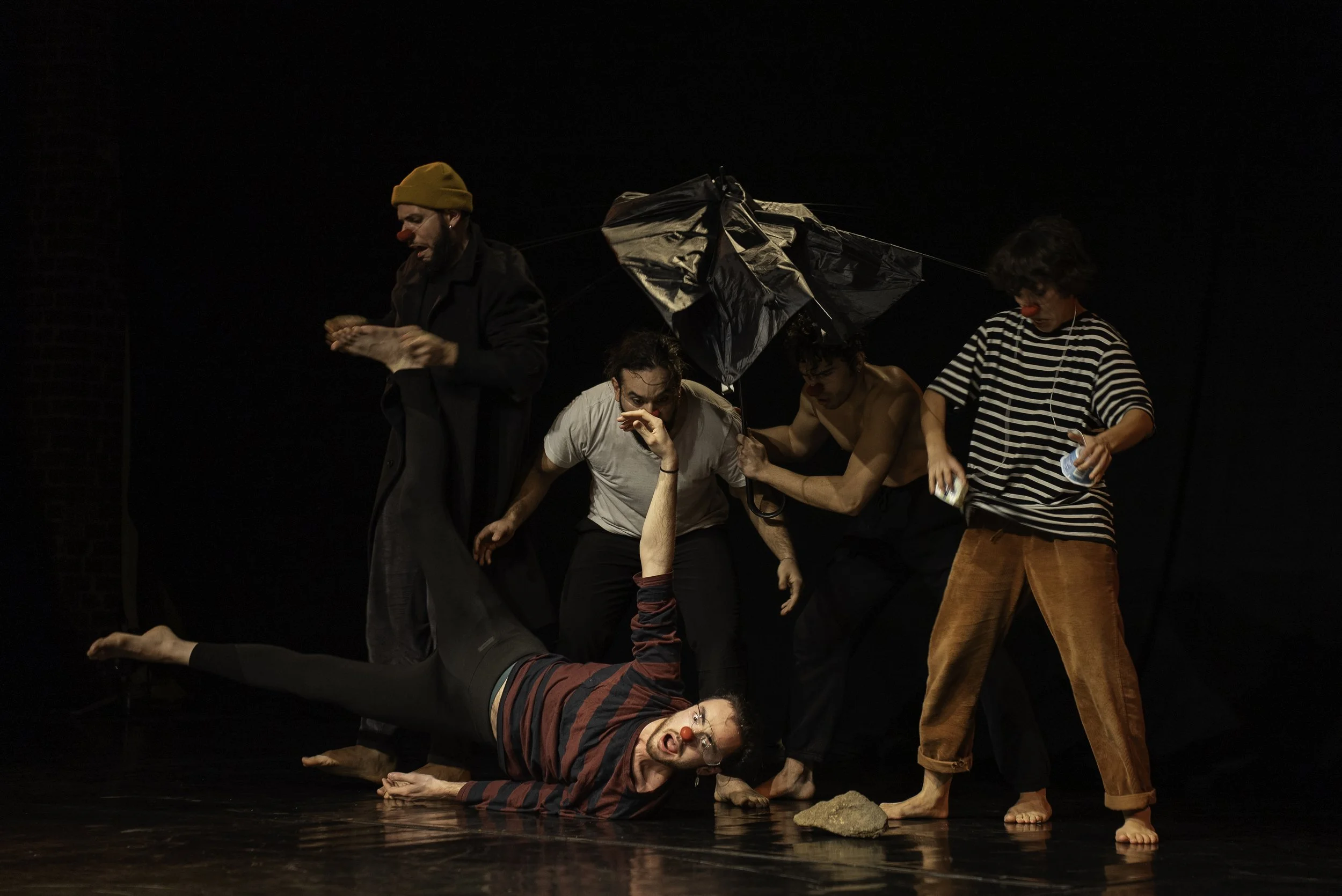 Five performers on stage with dark background, one lying on the floor with a clown nose, others standing or crouching, holding or interacting with various objects, including a black umbrella and a rock, during a theatrical performance.