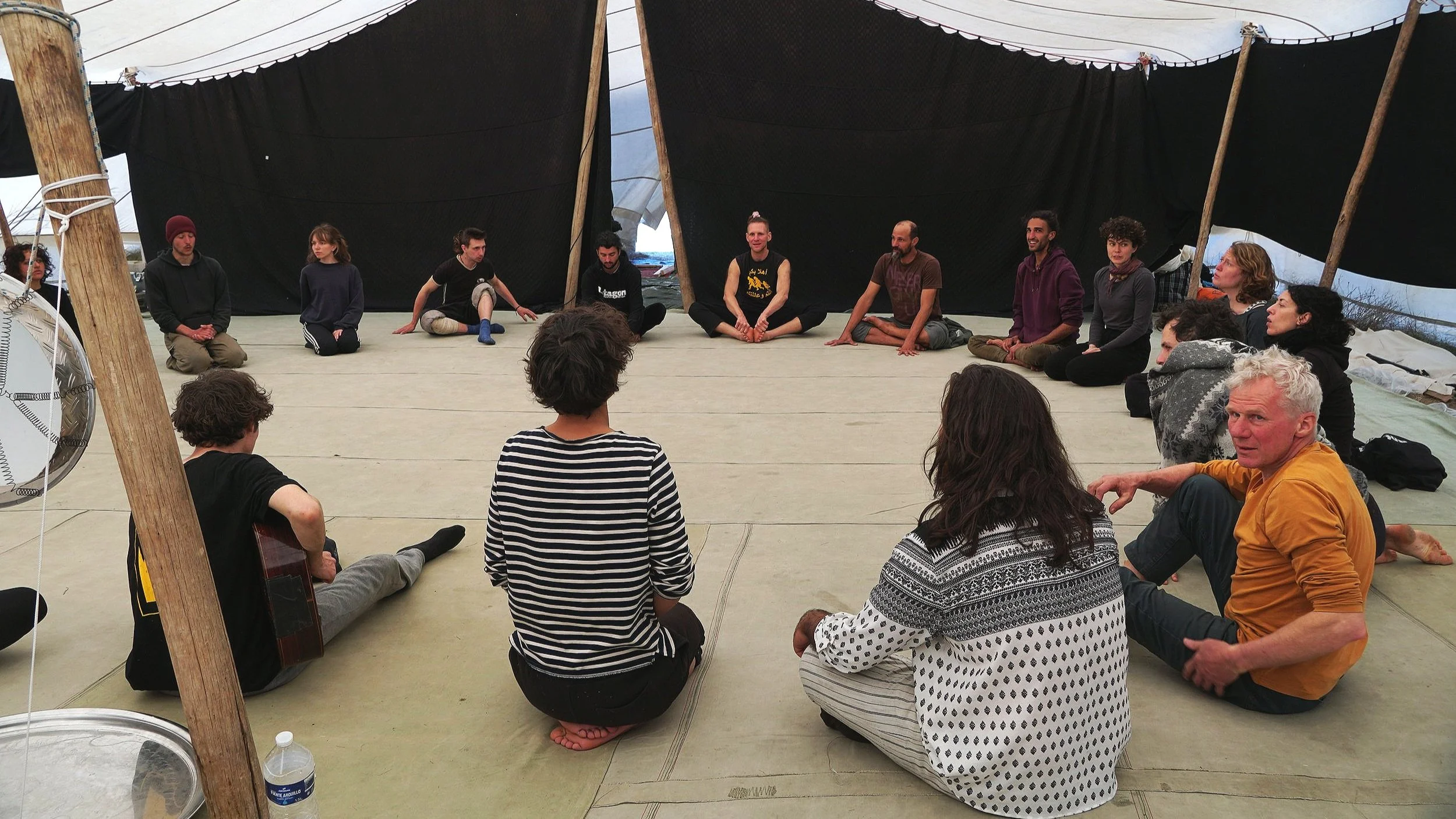 Group of people sitting in a circle on the floor inside a tent, engaged in a discussion or meditation, with black fabric walls and wooden support poles.