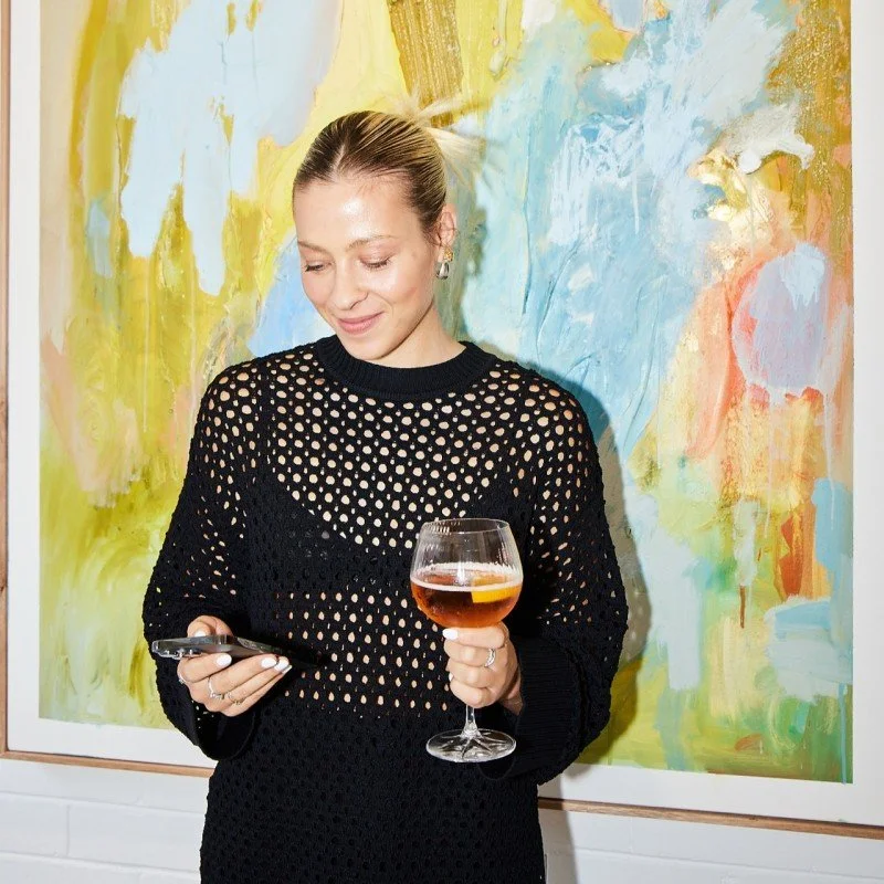 A young woman with hair in a bun, looking down at her ohone, while holding a drink, standing in front of an art piece