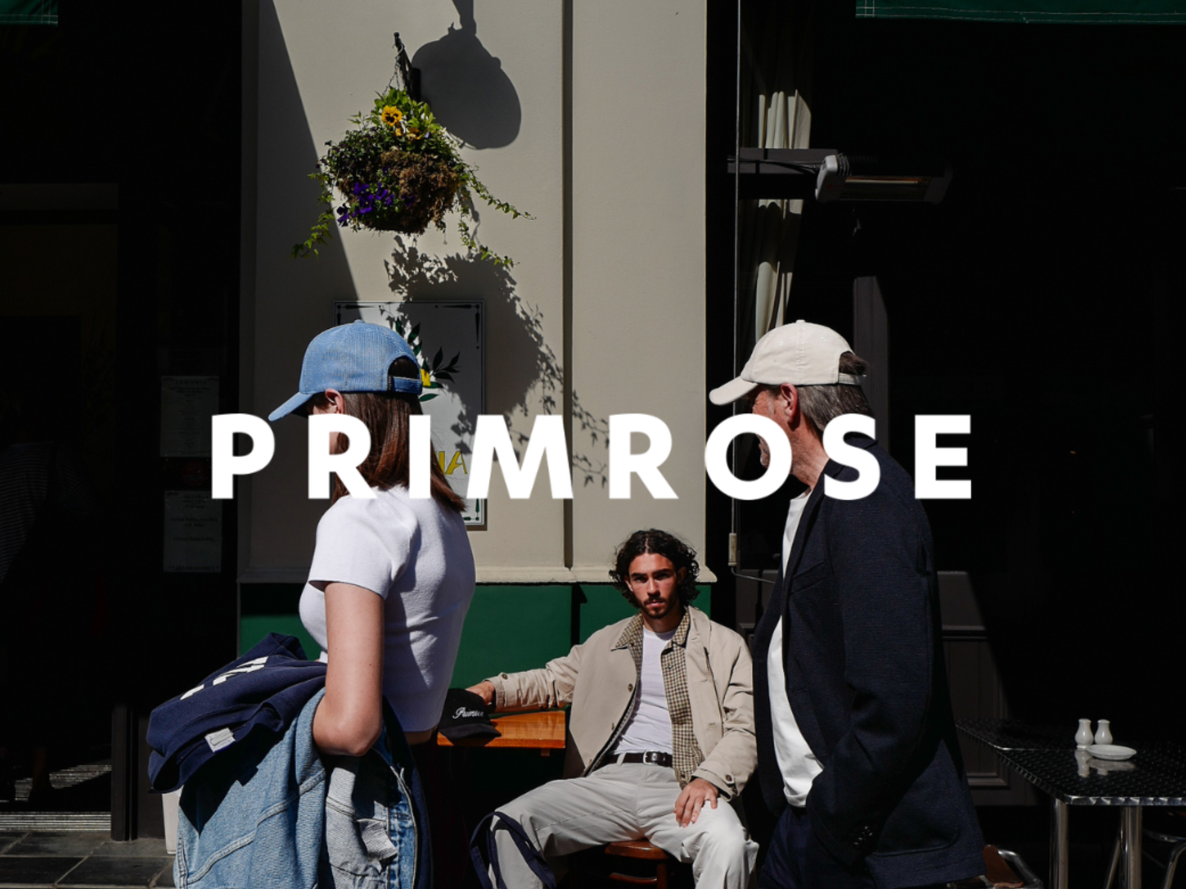Image of man sitting outside cafe staring at camera and two people in caps walking past. Primrose Logo is overlayed